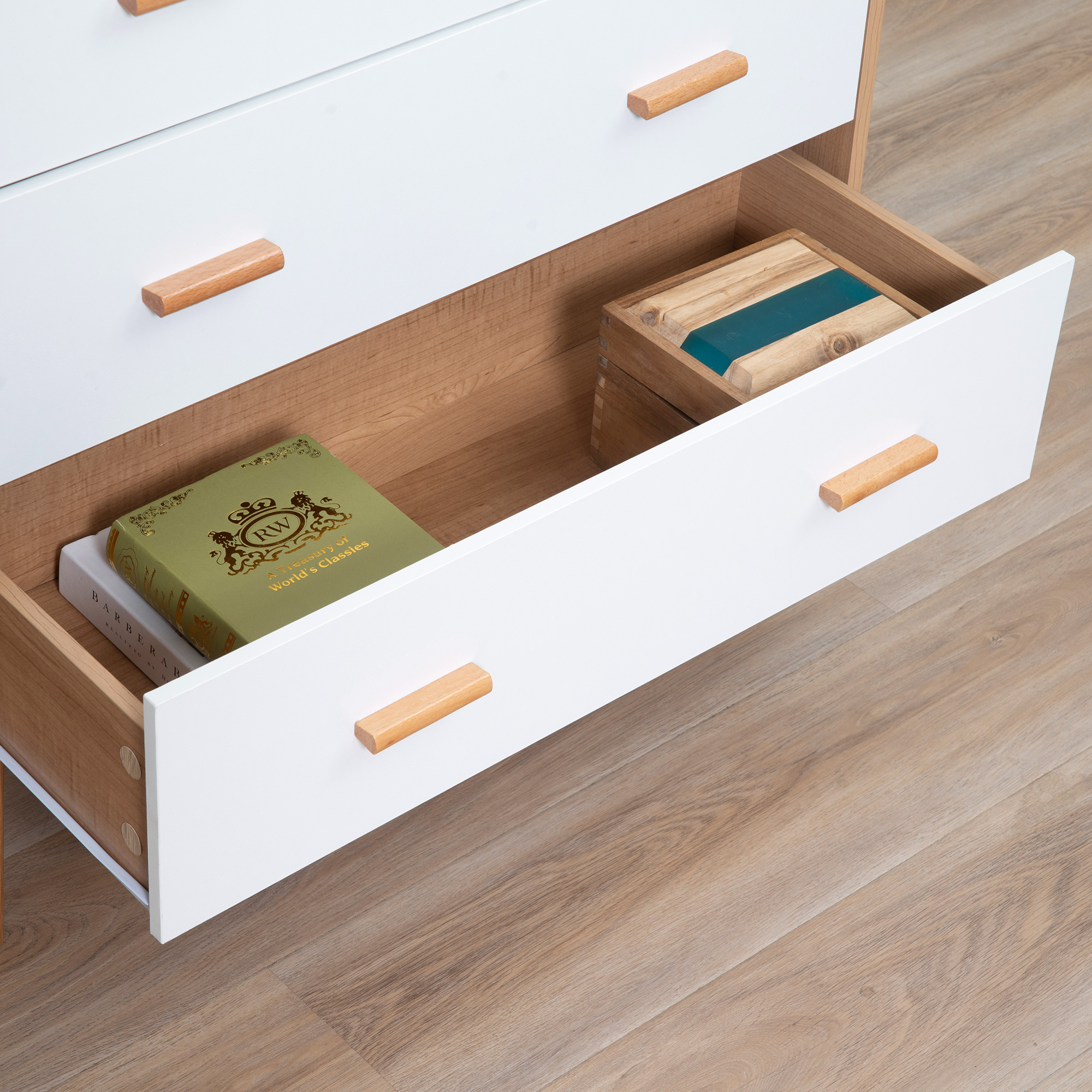 Una cómoda blanca con tiradores de madera. Un cajón está abierto, mostrando libros y una caja de madera.