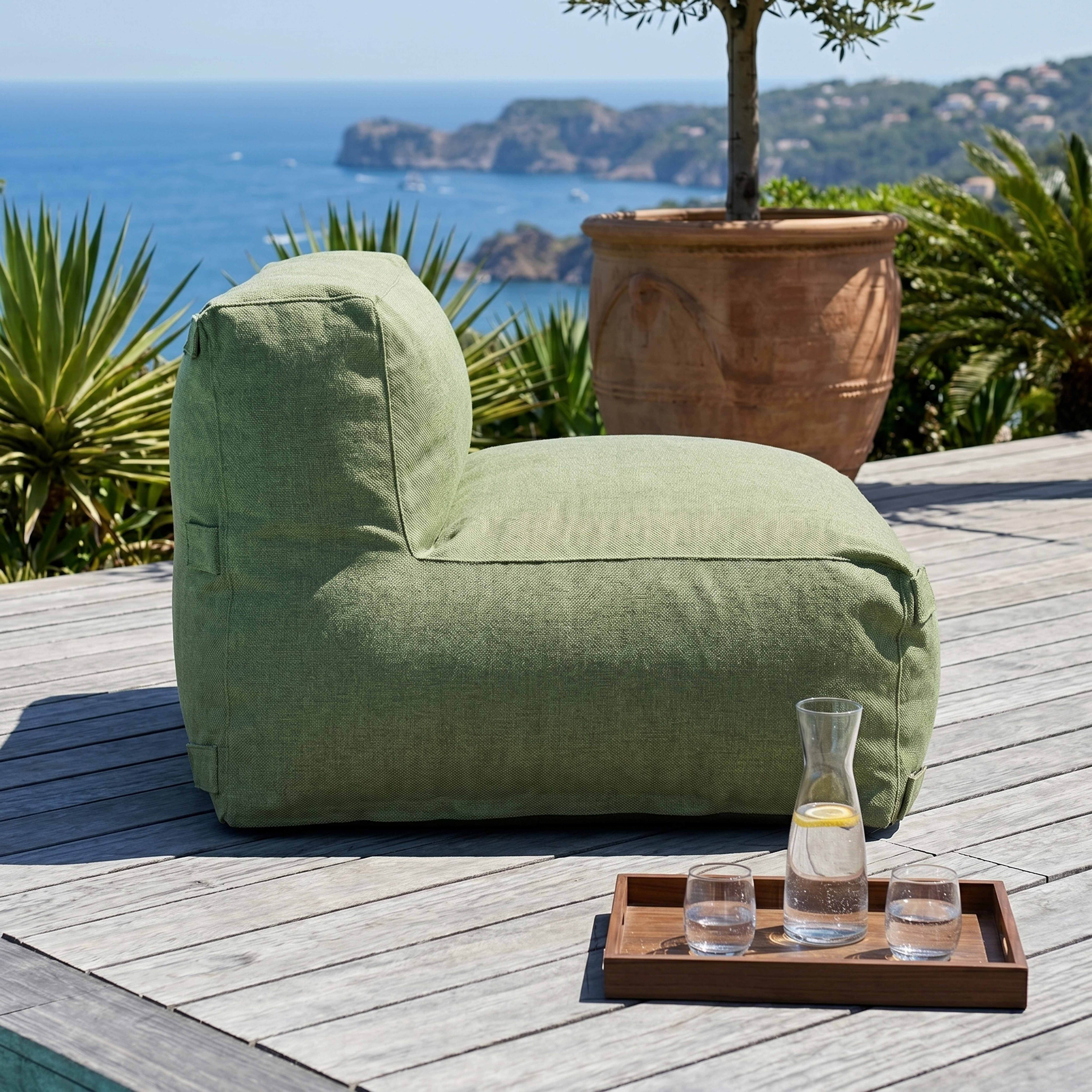 Ein grüner Sitzsack steht auf einer Holzterrasse am Meer mit Gläsern und Karaffe.