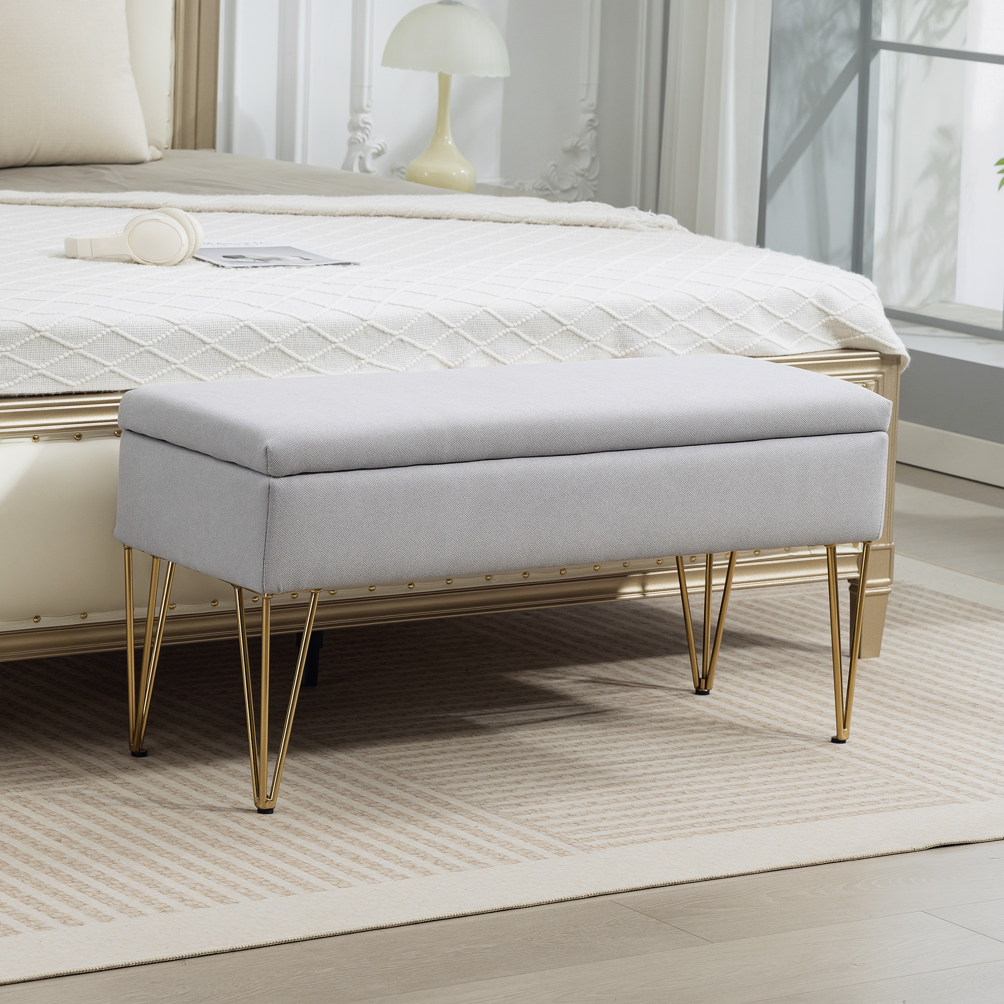 Panca contenitore grigia con gambe dorate davanti a un letto con copriletto bianco.