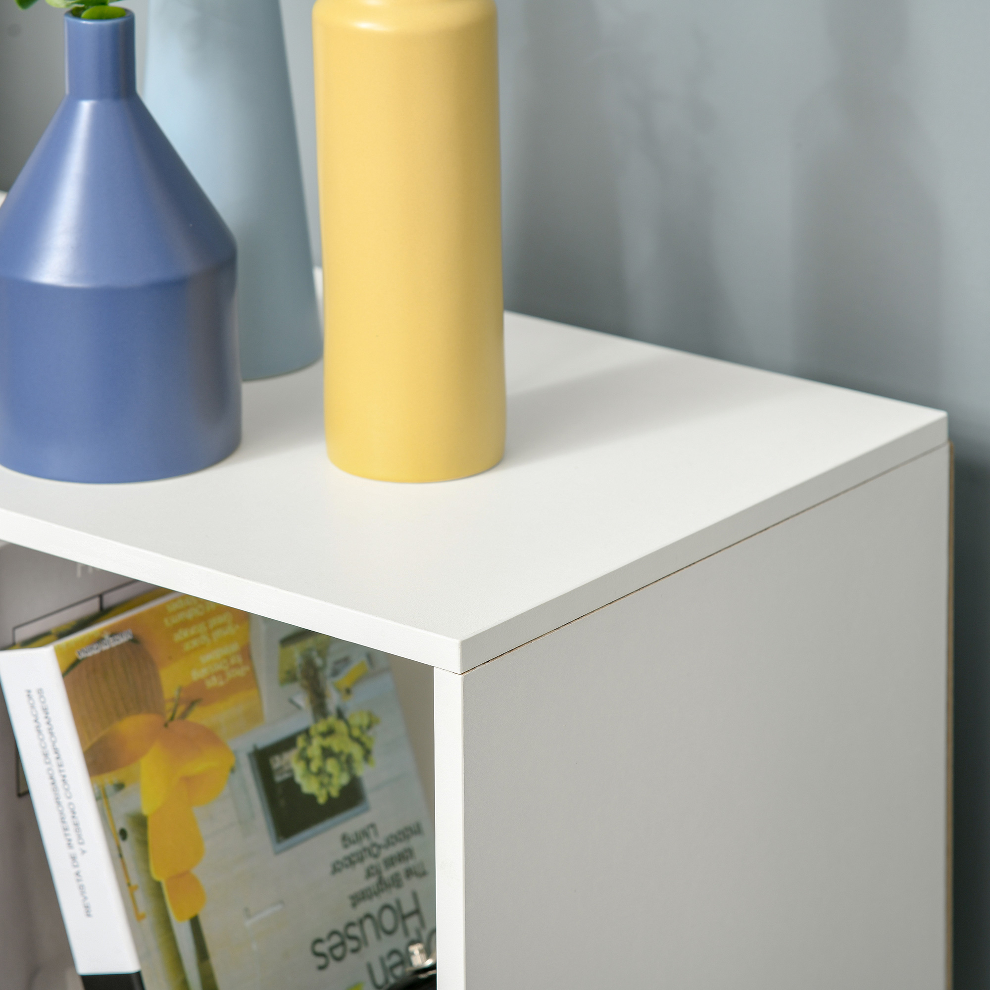 Un jarrón azul, uno celeste y uno amarillo sobre un estante blanco. Revistas debajo.