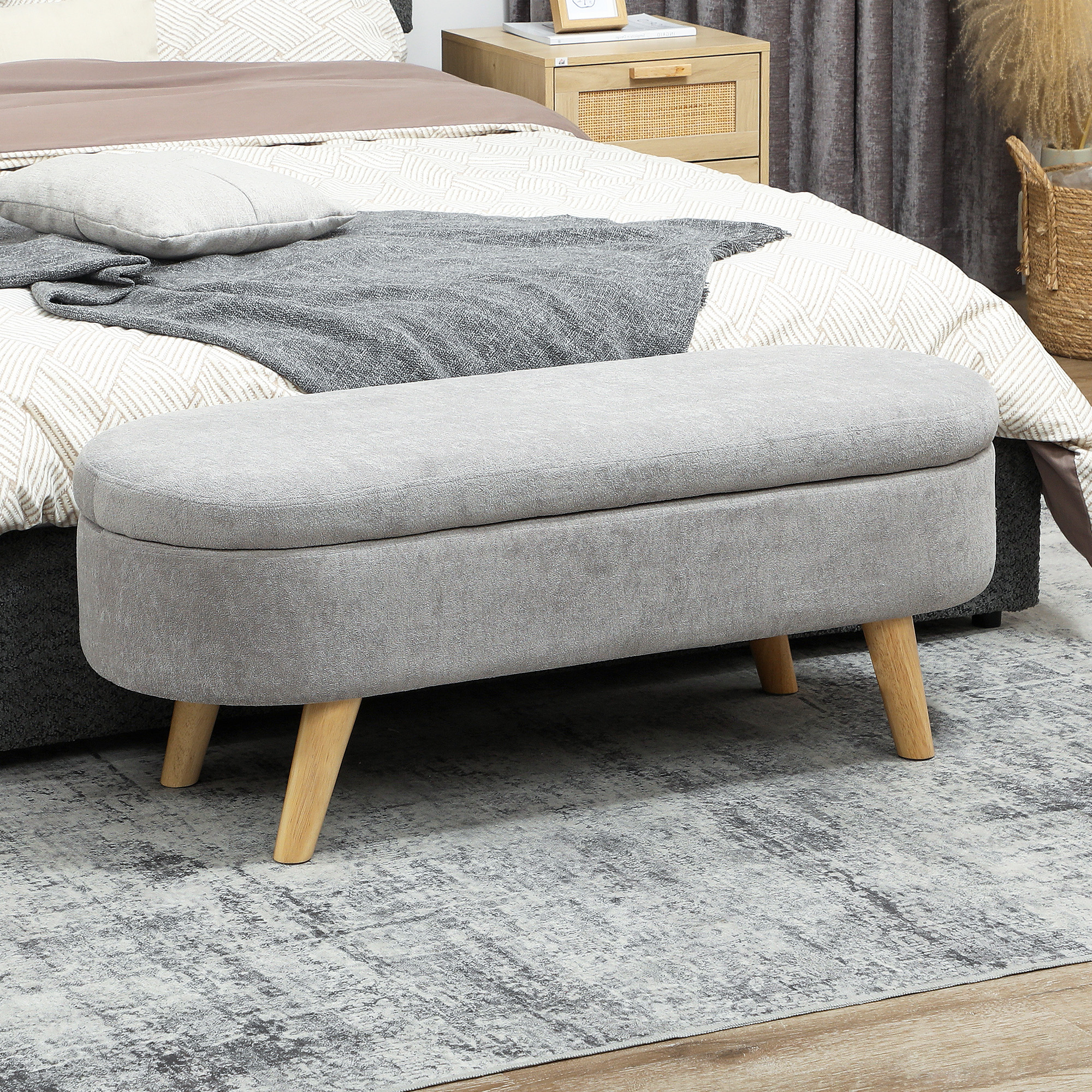 Un otomano gris con patas de madera delante de una cama con manta gris.