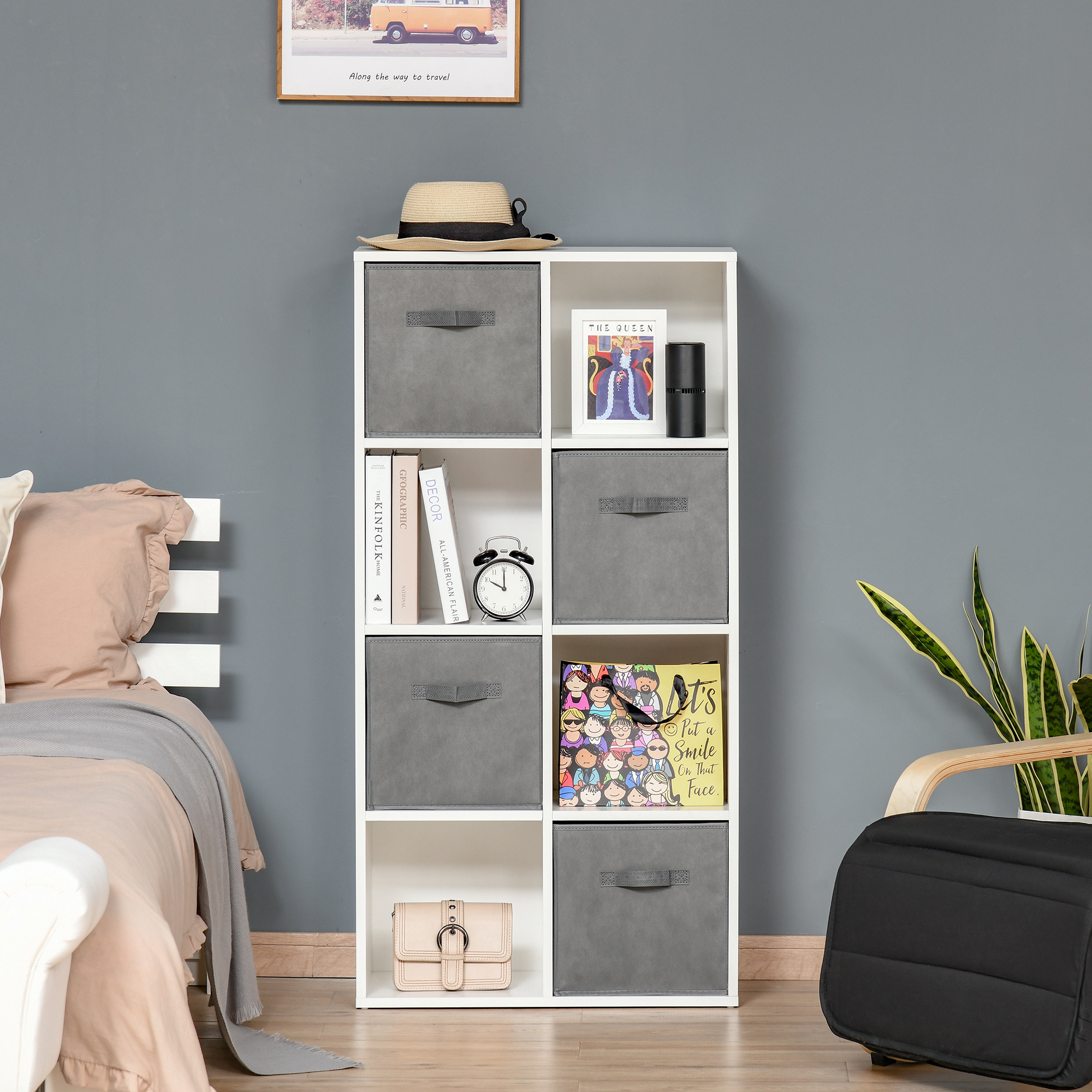 Un estante cúbico blanco tiene libros, cubos, un cuadro y un bolso. Una cama está a la izquierda.