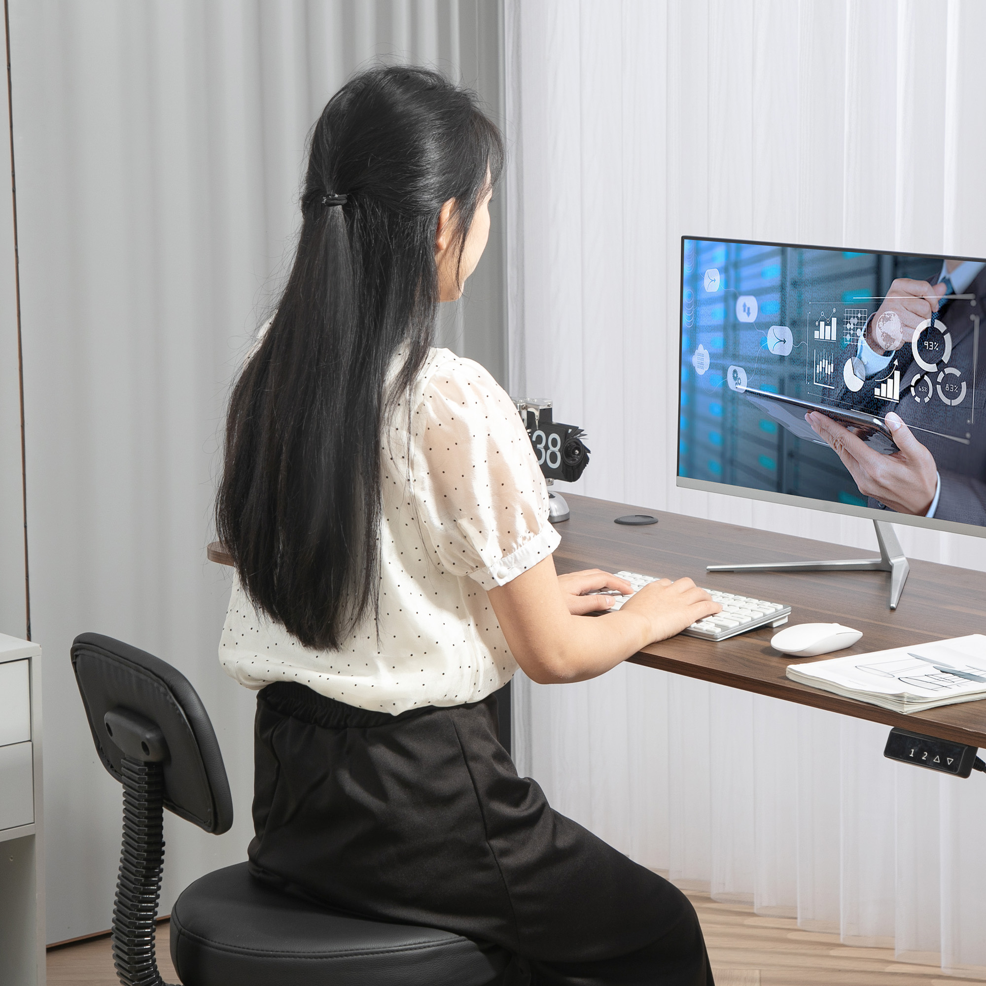 Mujer en escritorio teclea en teclado frente a pantalla con datos.