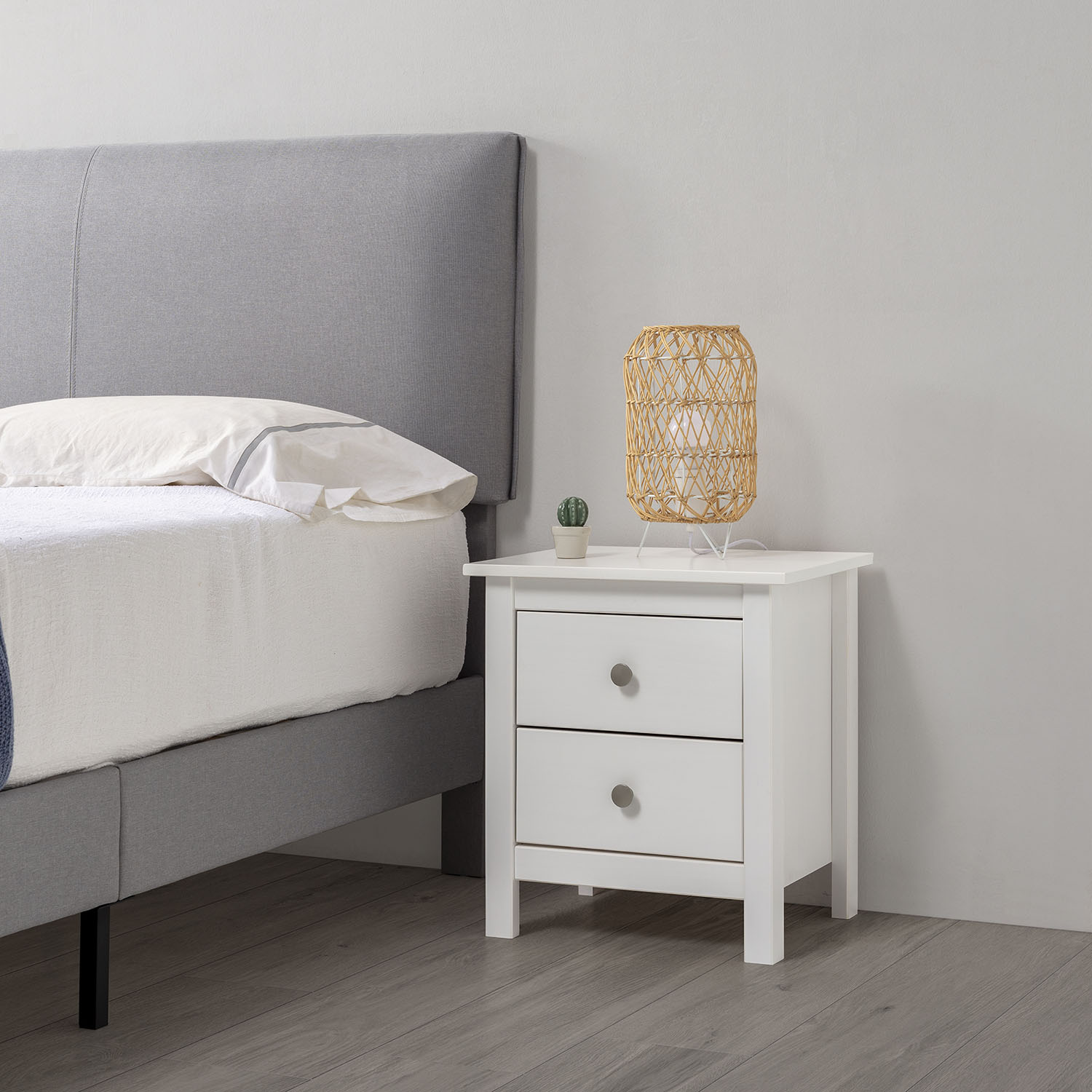 Cama con cabecero tapizado gris, mesita de noche blanca, lámpara y cactus.