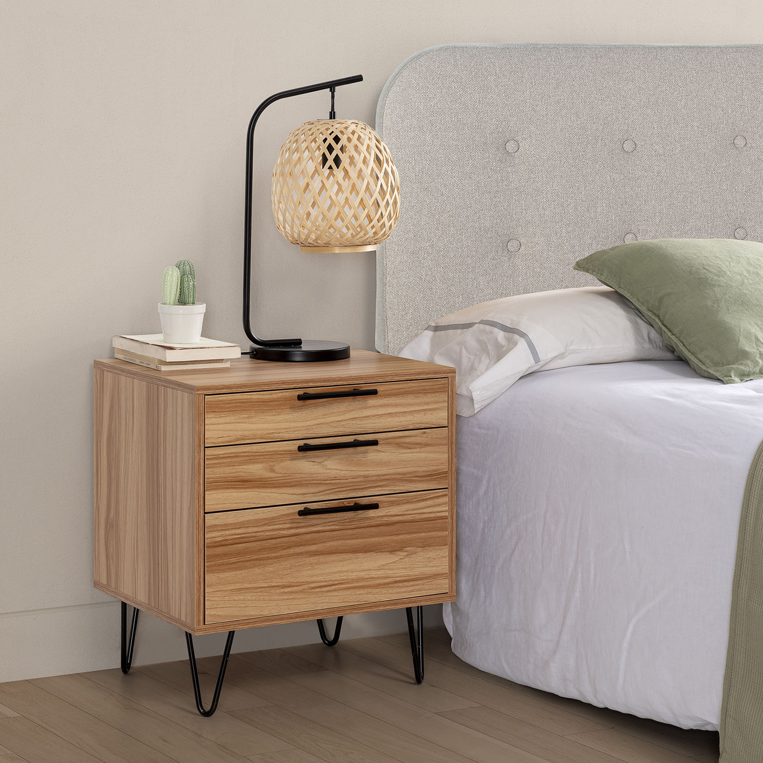 Mesita de noche de madera con tres cajones, lámpara, libros y cactus junto a cama.