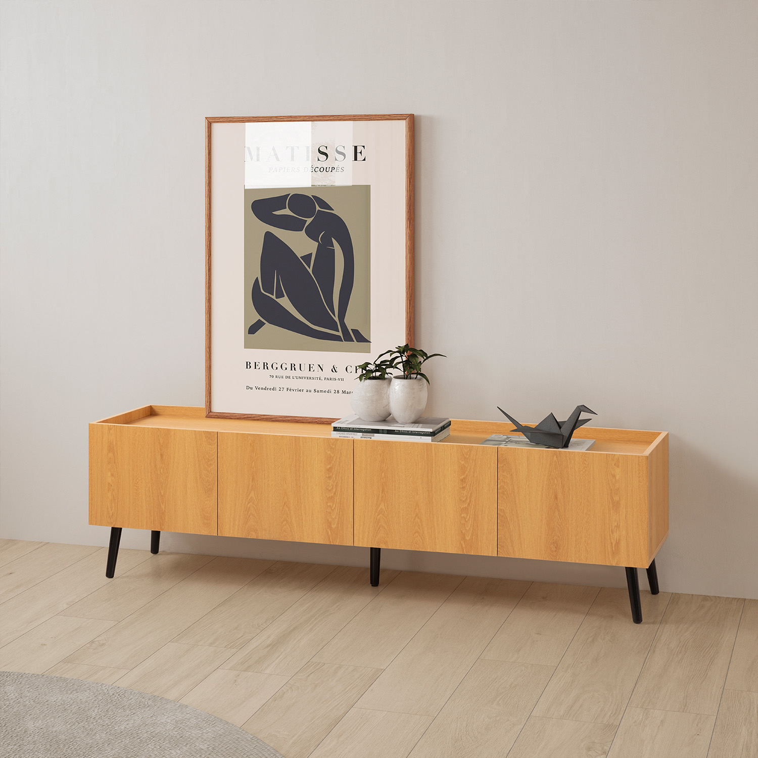 Un póster de arte de Matisse cuelga sobre un mueble de madera con patas negras. Dos plantas y una grulla de papel encima.