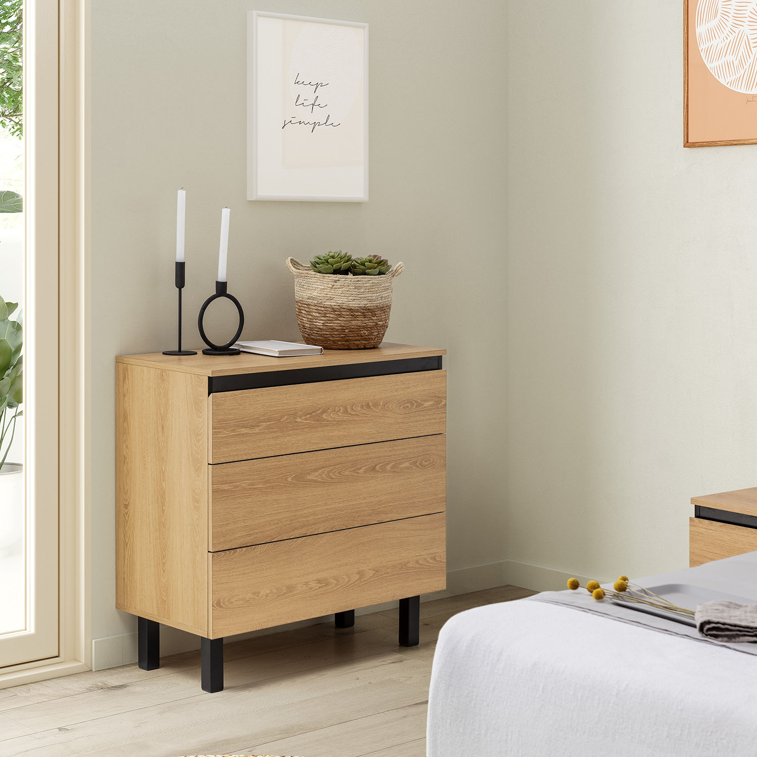 Cómoda de madera con tres cajones, velas y planta. Un cuadro colgado en la pared.
