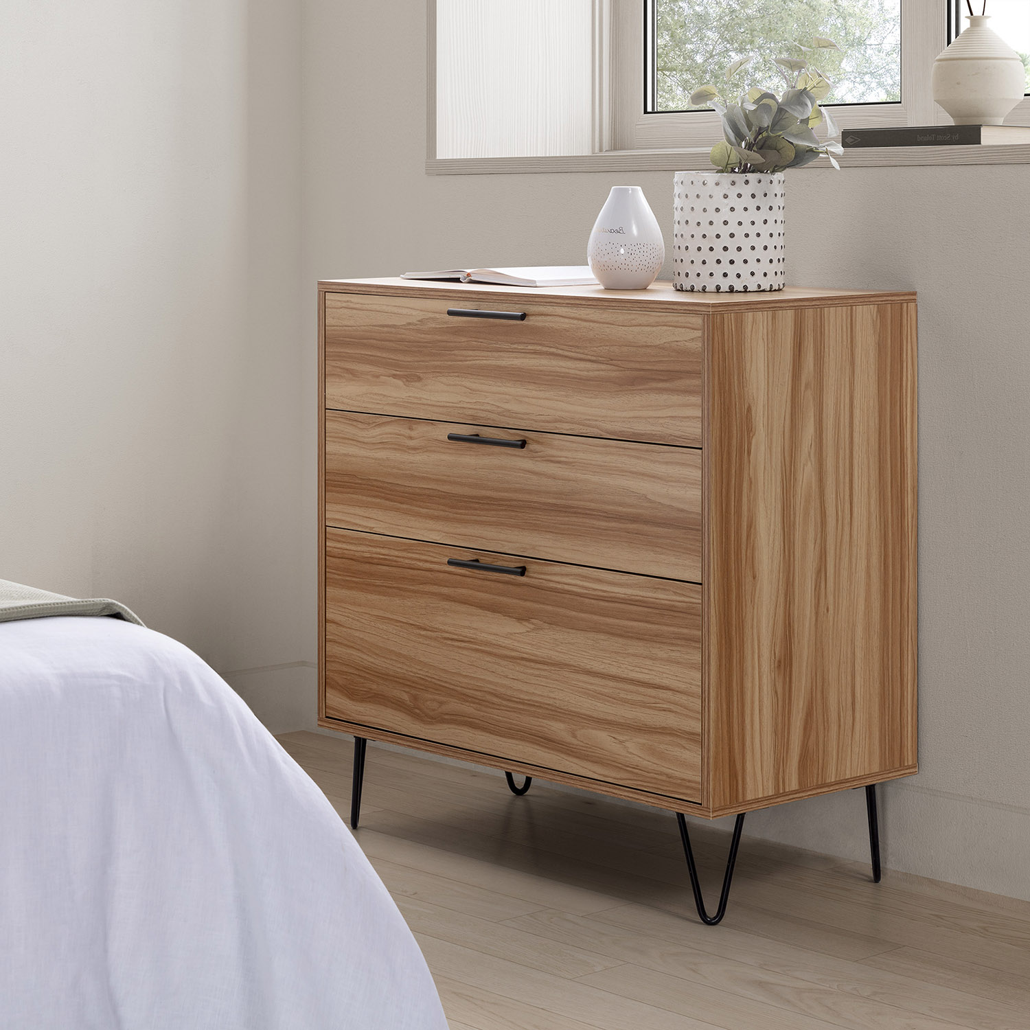 Cómoda de madera con tres cajones y patas negras, junto a una cama.