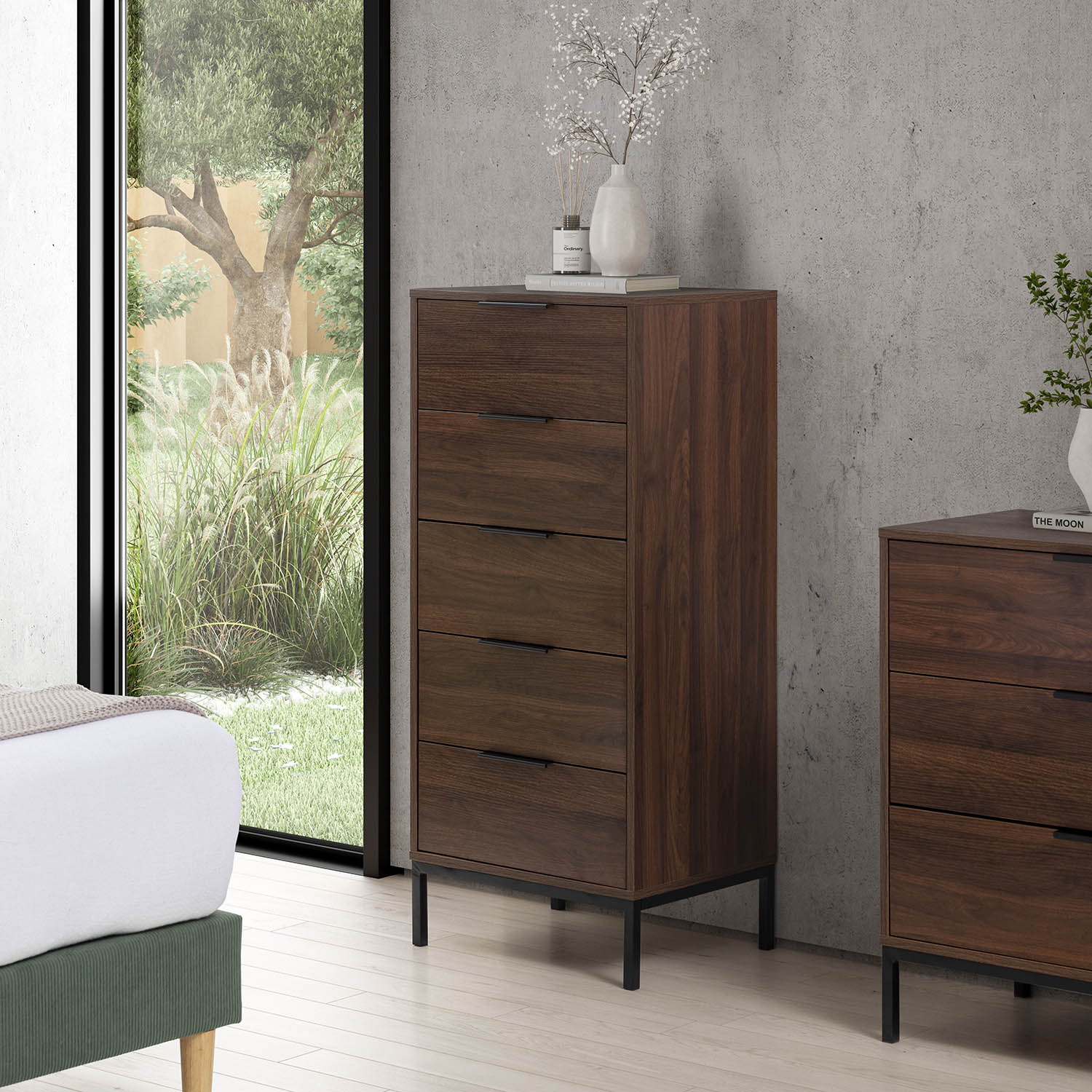 Una cómoda de madera marrón de cinco cajones se encuentra junto a una cama y una ventana.