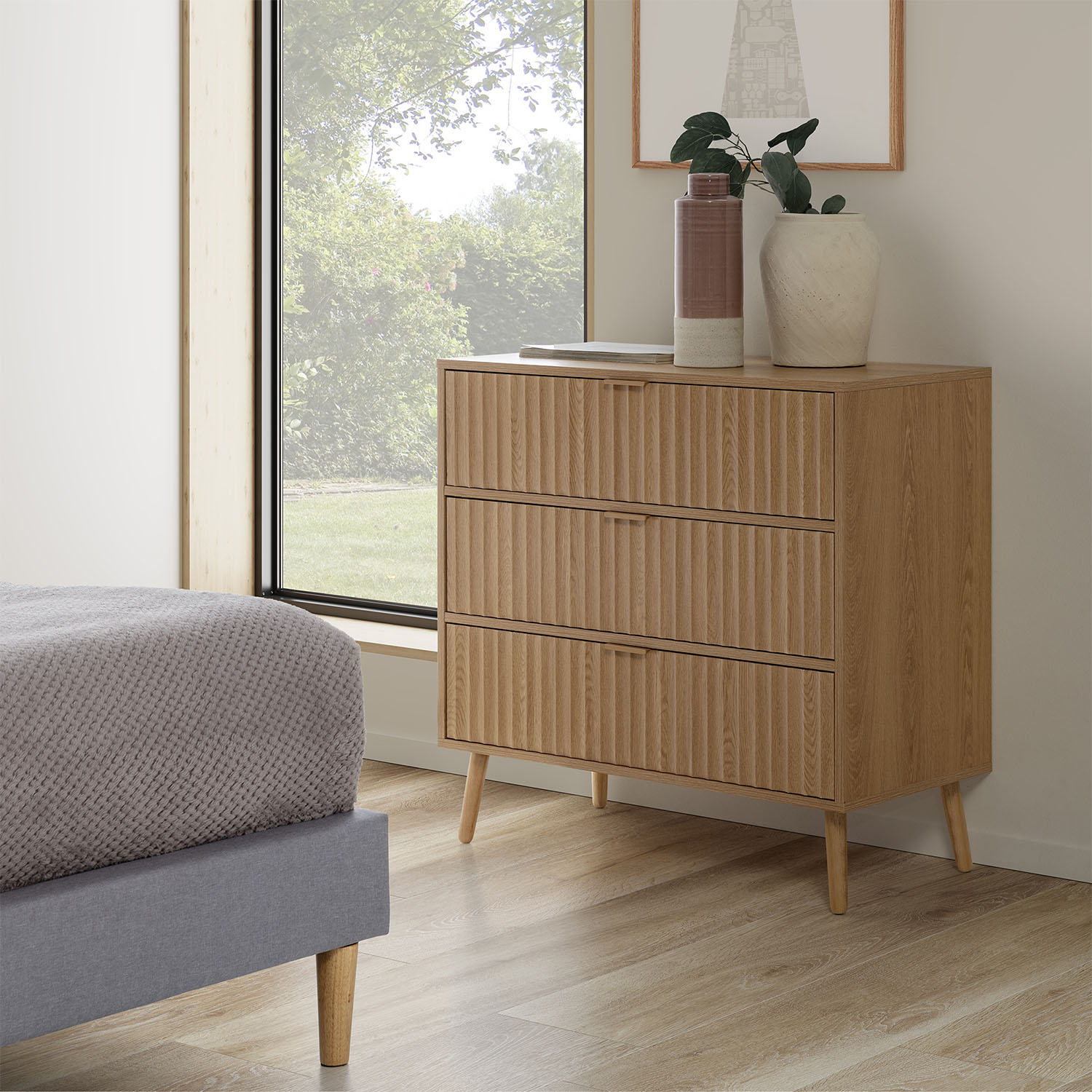 Una cómoda de madera con tres cajones se encuentra junto a una cama con manta gris.