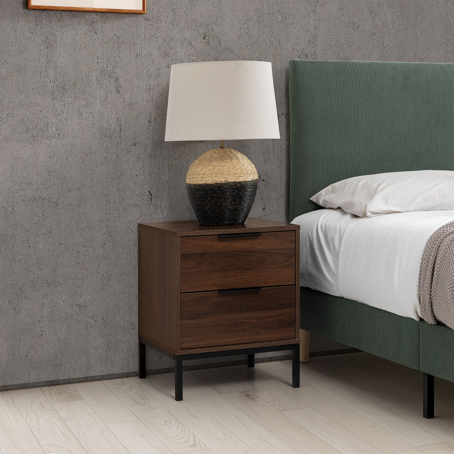 Una mesita de noche de madera con una lámpara junto a una cama verde sobre suelo de madera.