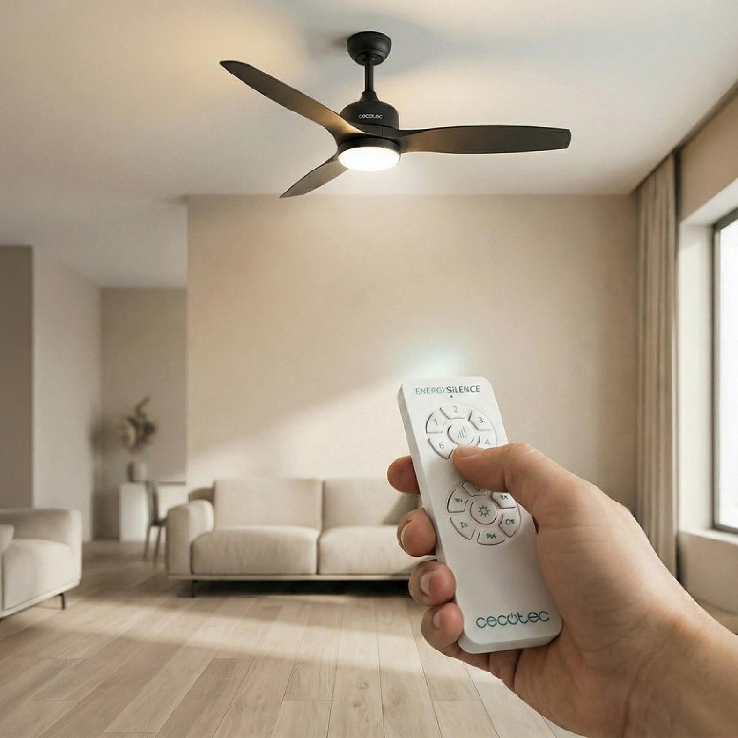 Una mano tiene un telecomando bianco davanti a un ventilatore da soffitto con luce in un soggiorno.