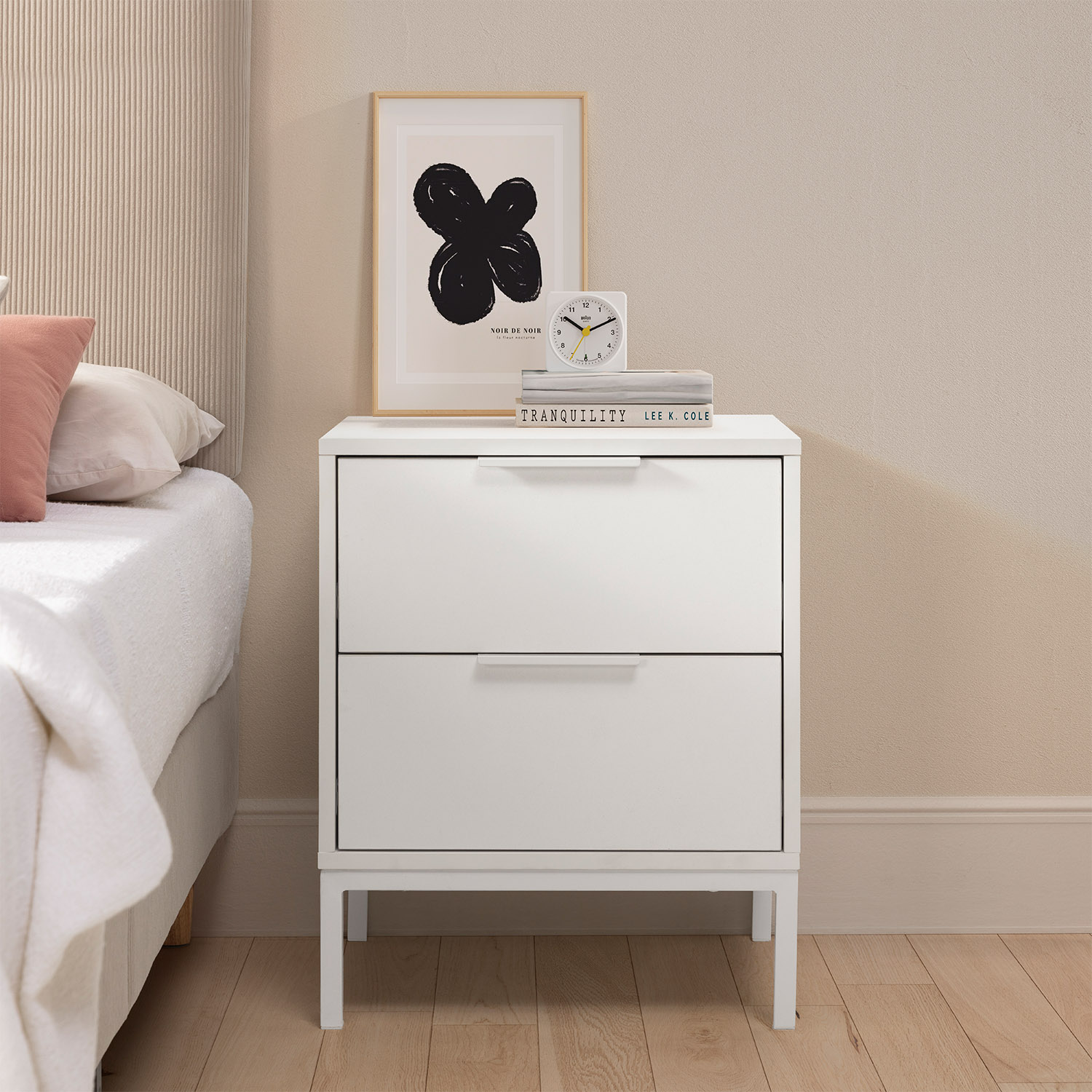 Una mesita de noche blanca de dos cajones junto a una cama con almohadas y manta.
