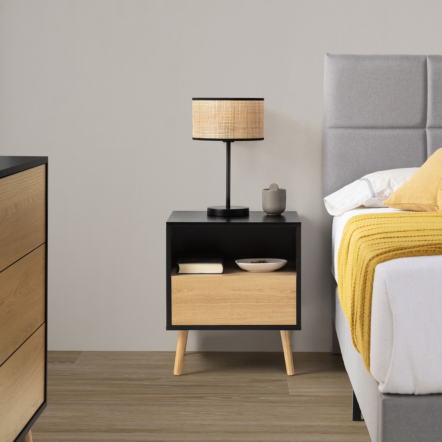 Una mesita de noche negra y de madera con lámpara, libro y cuenco junto a una cama.