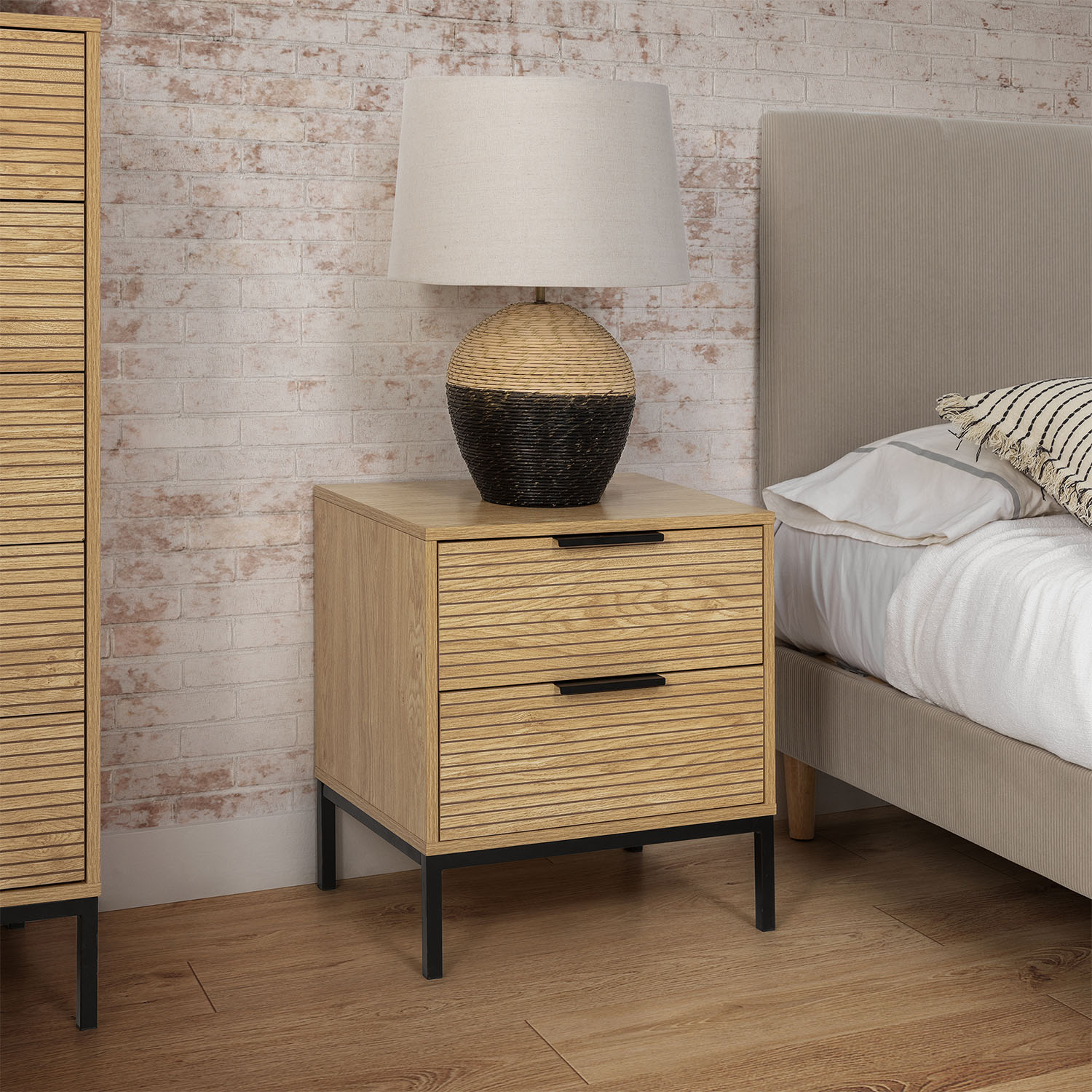 Mesita de noche de madera con dos cajones junto a una cama con cabecero beige.