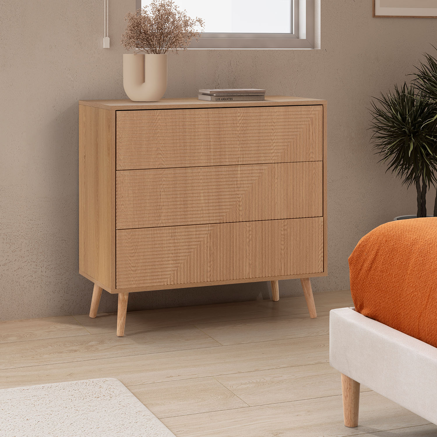 Una cómoda de madera con tres cajones se apoya en cuatro patas junto a una ventana.