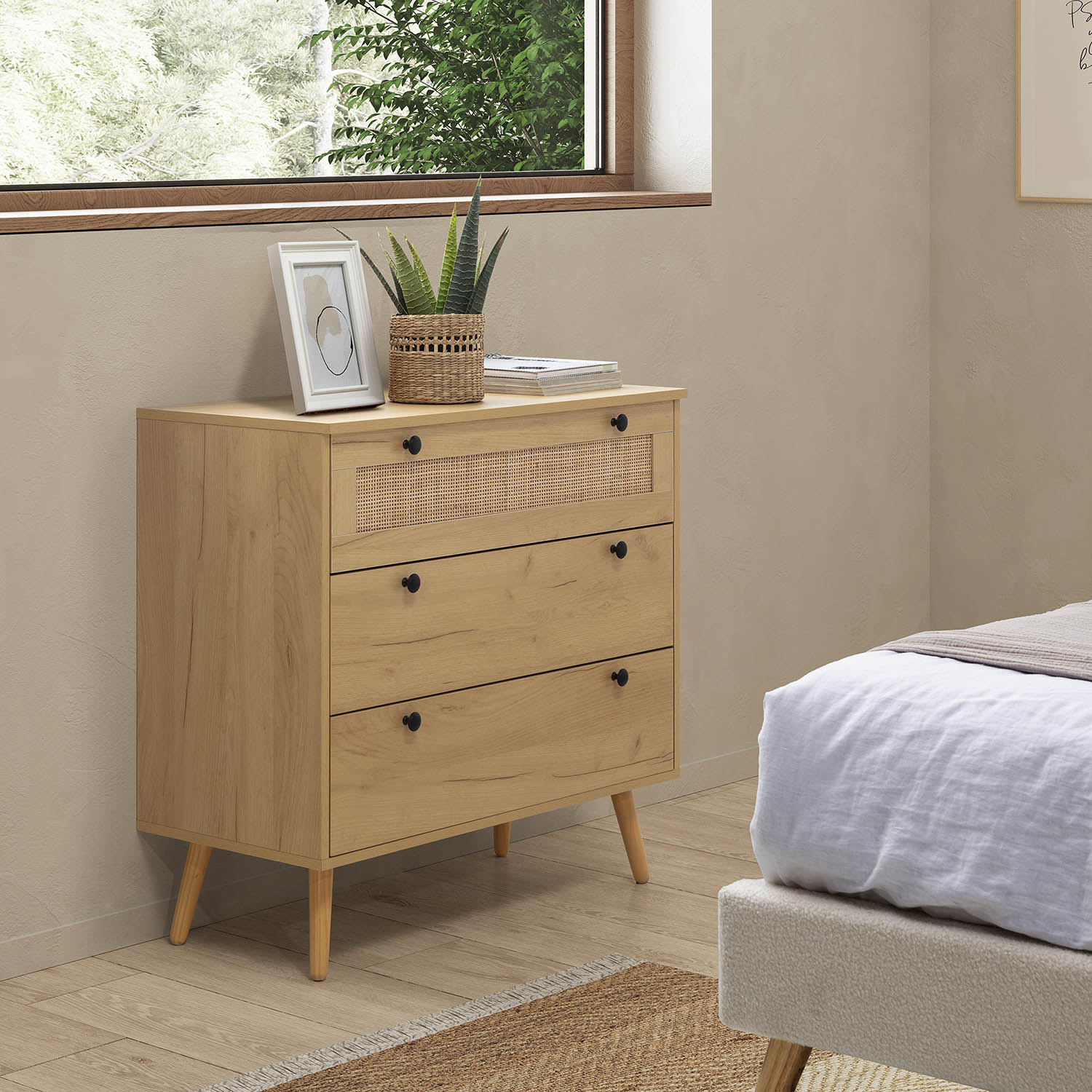 Cómoda de madera con frente de ratán junto a una cama, ventana con vegetación.