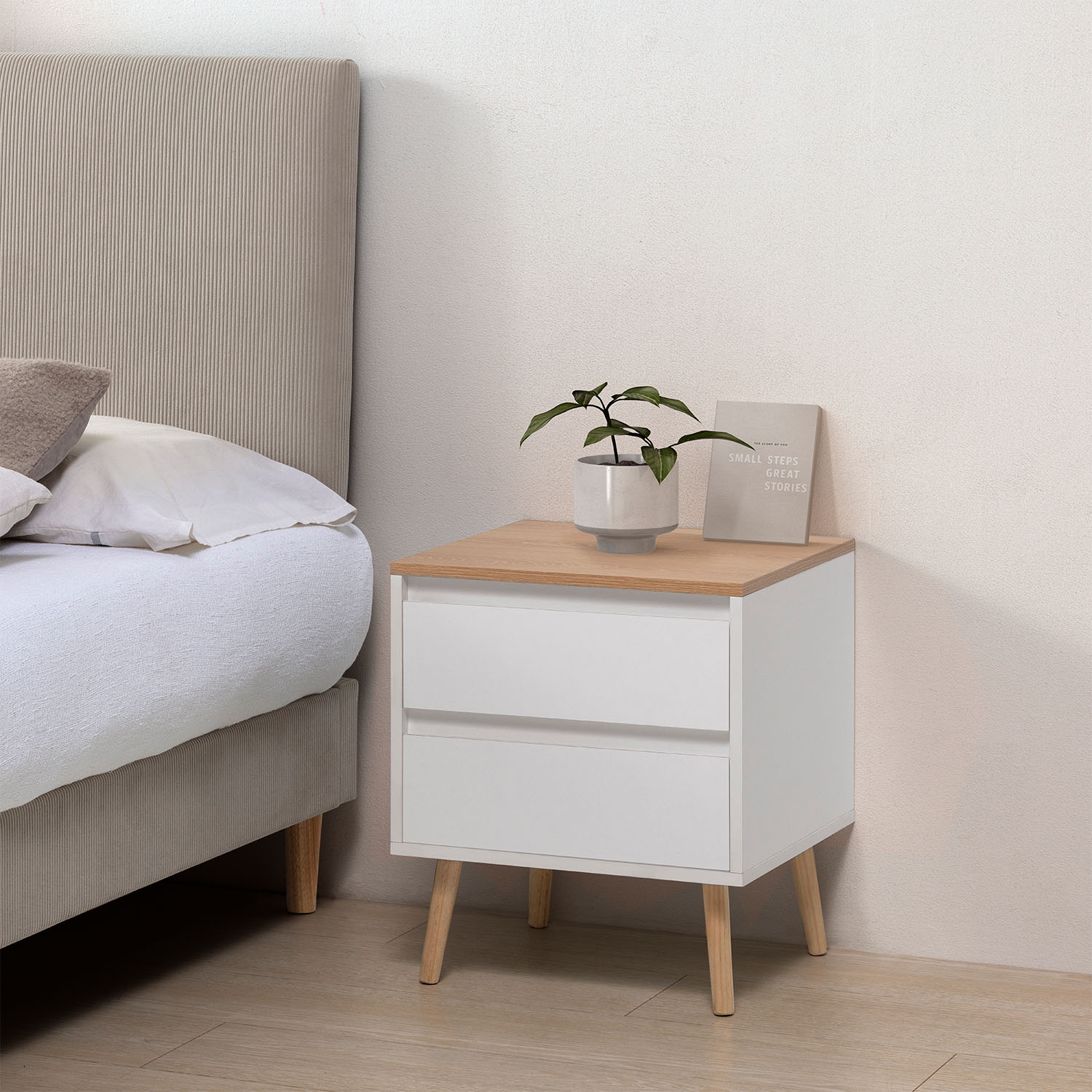Una mesita de noche blanca y de madera junto a una cama con cabecero de tela.