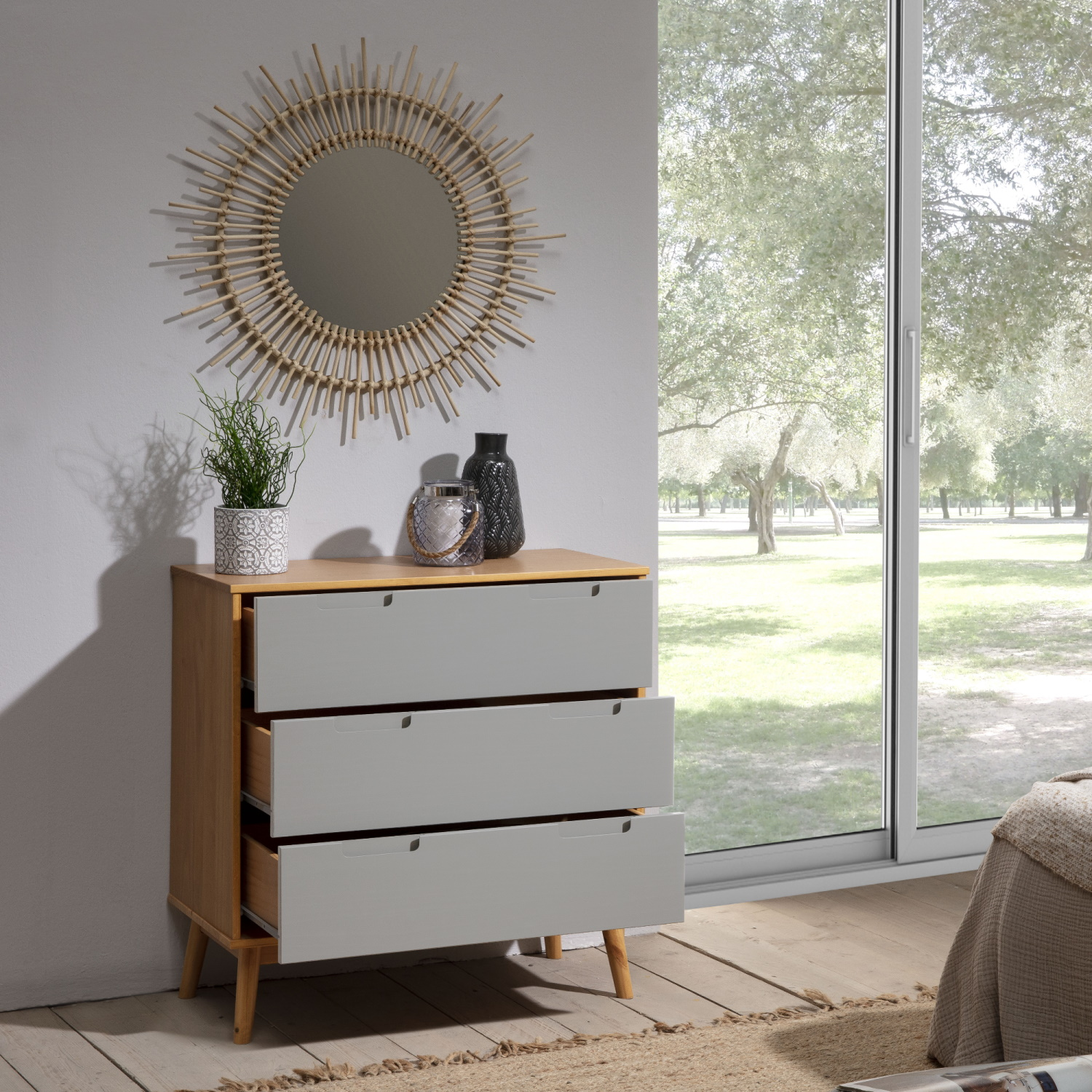 Cómoda de madera con tres cajones junto a una ventana con vistas a la vegetación.