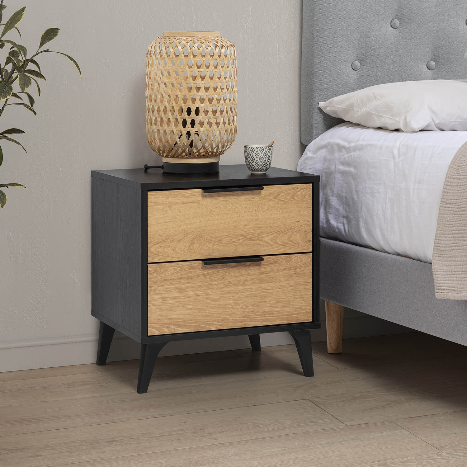 Mesita de noche con cajones de madera y marco negro junto a una cama.