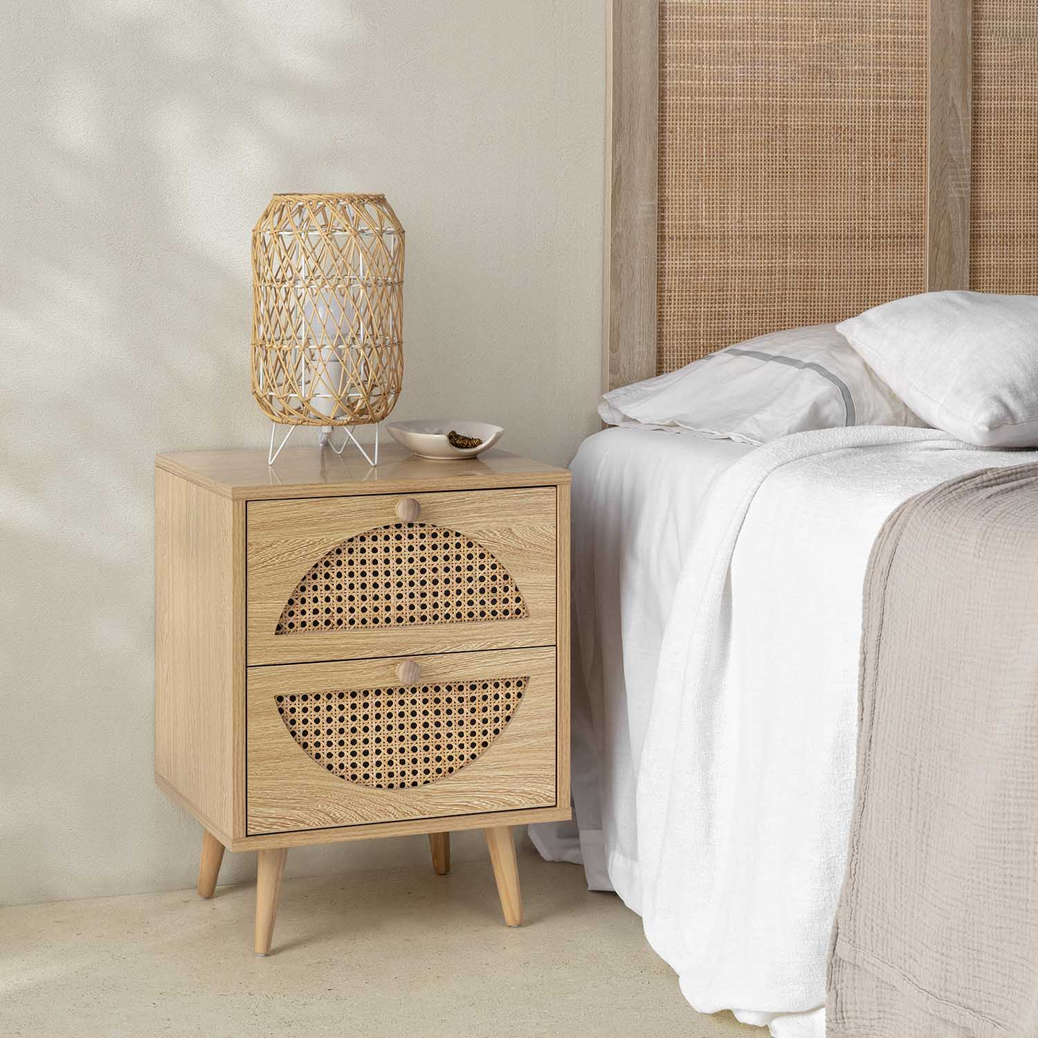 Una mesita de noche de madera con detalles de ratán junto a una cama con ropa de cama blanca.