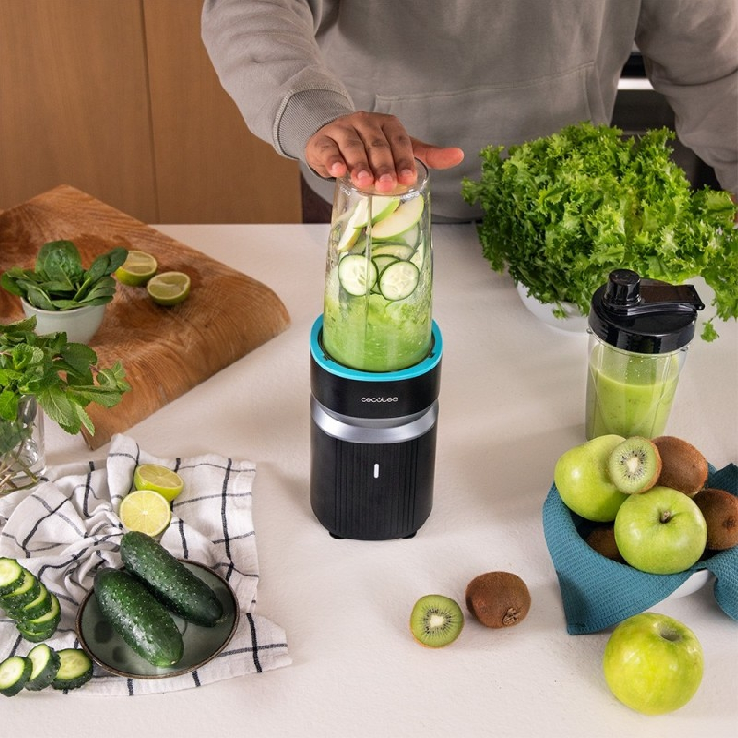 Persona che prepara un frullato verde con un frullatore, circondata da frutta e verdura.