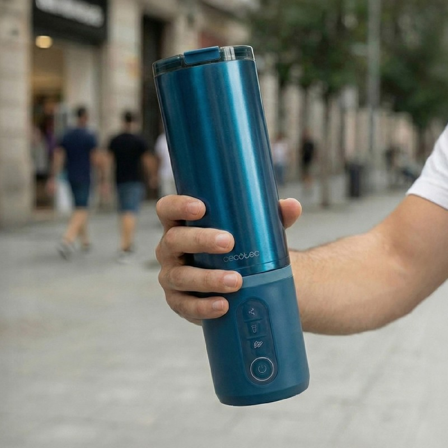 Una persona sostiene una licuadora portátil azul al aire libre. La licuadora tiene un acabado metálico y una base azul.