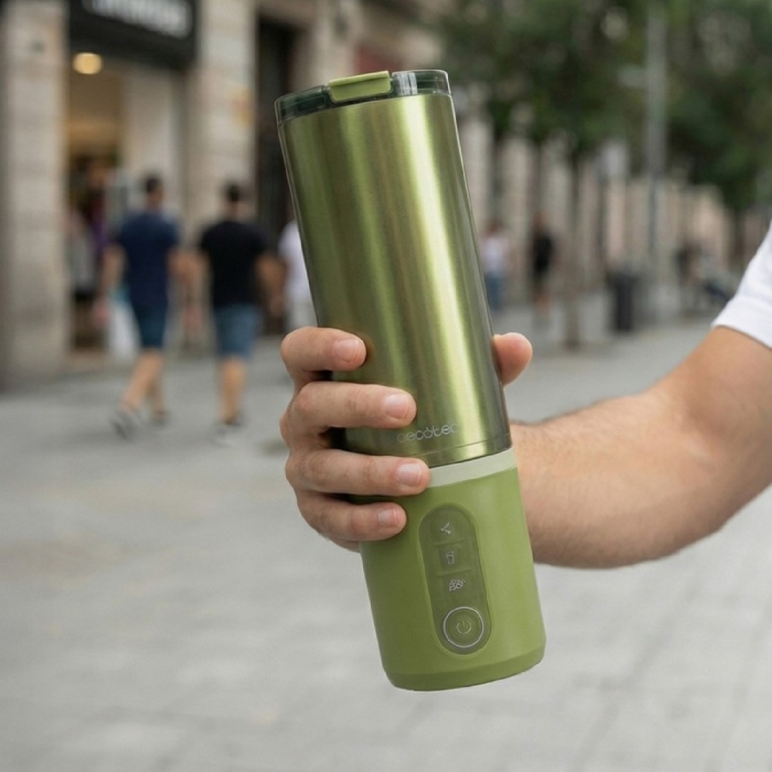 Batidora portátil verde sostenida en mano en una calle con gente desenfocada.