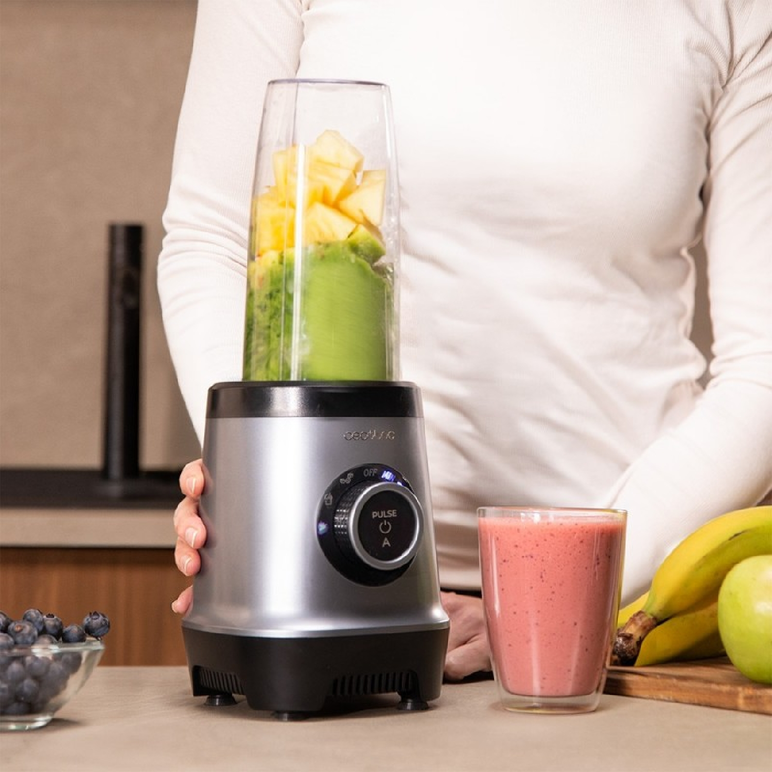 Una licuadora con frutas, un vaso de batido, plátanos y arándanos en una mesa.