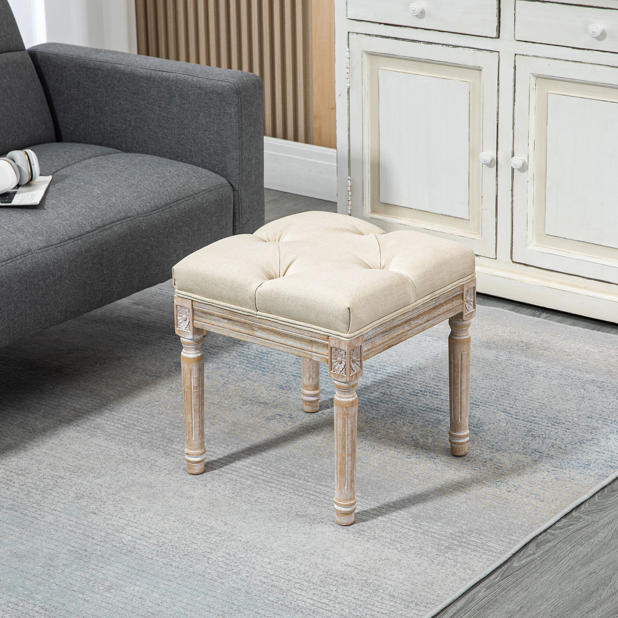 Un otomana beige con botones y patas de madera tallada se sienta sobre una alfombra.