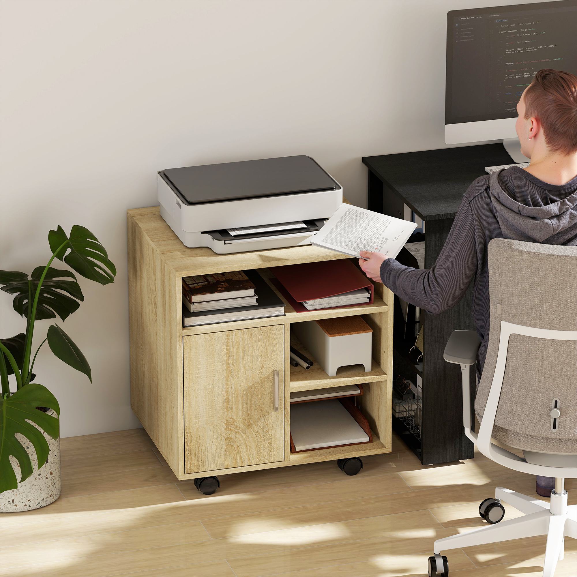 Un hombre se sienta frente a una computadora. A su lado, una impresora en un mueble con ruedas.