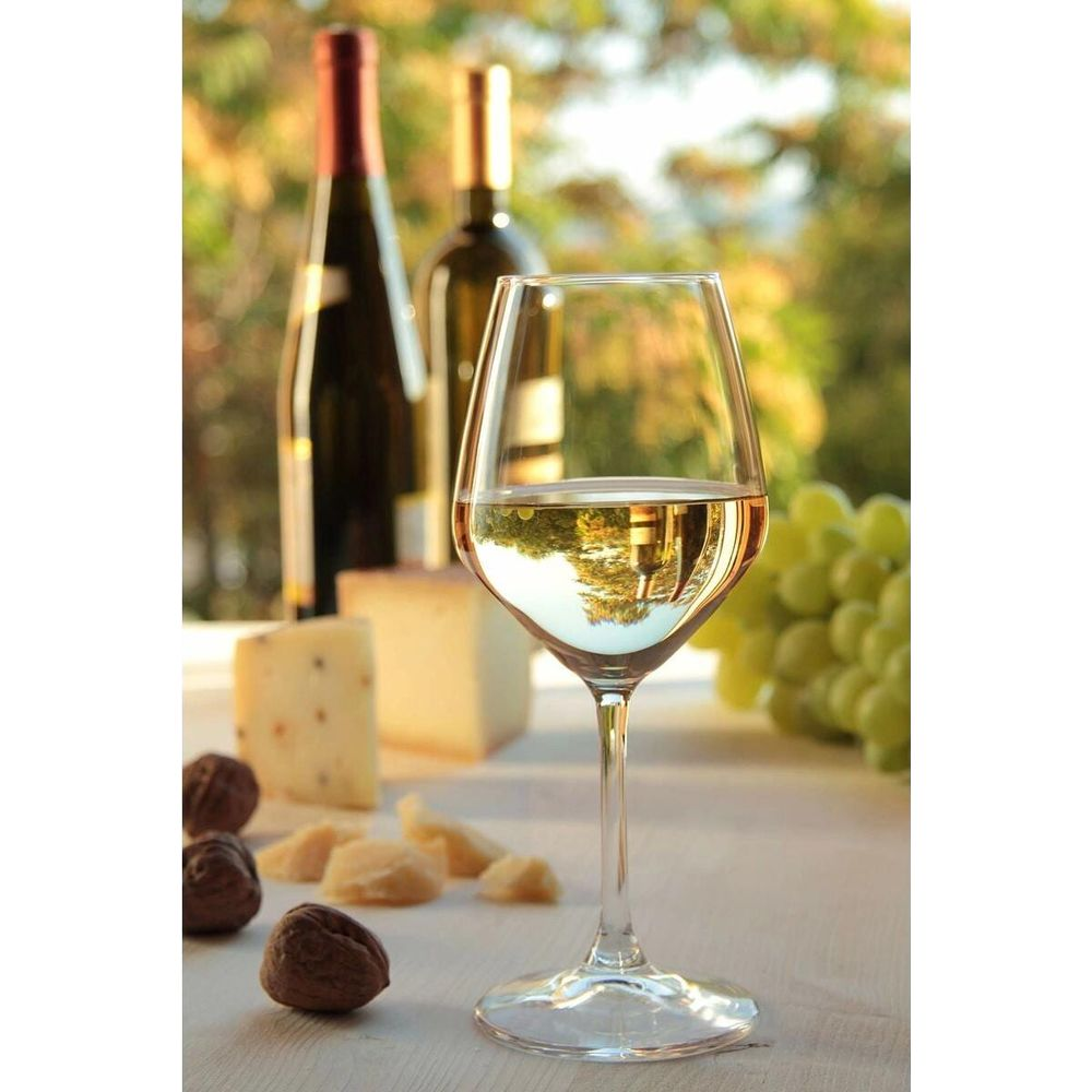 A glass of white wine sits on a table with cheese, walnuts, and grapes. Two bottles of wine stand behind.