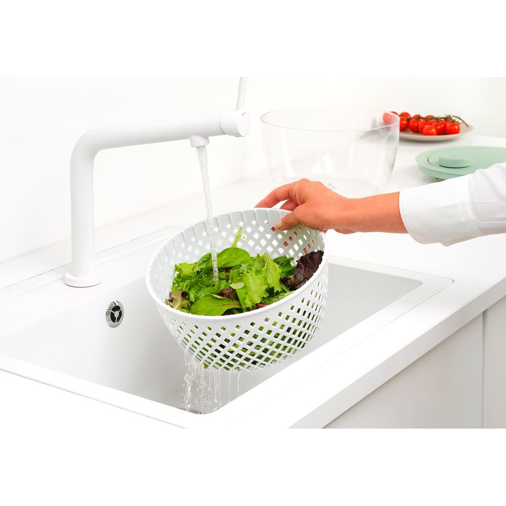Una mano tiene una centrifuga per insalata bianca con lattuga sotto l'acqua corrente in un lavandino.
