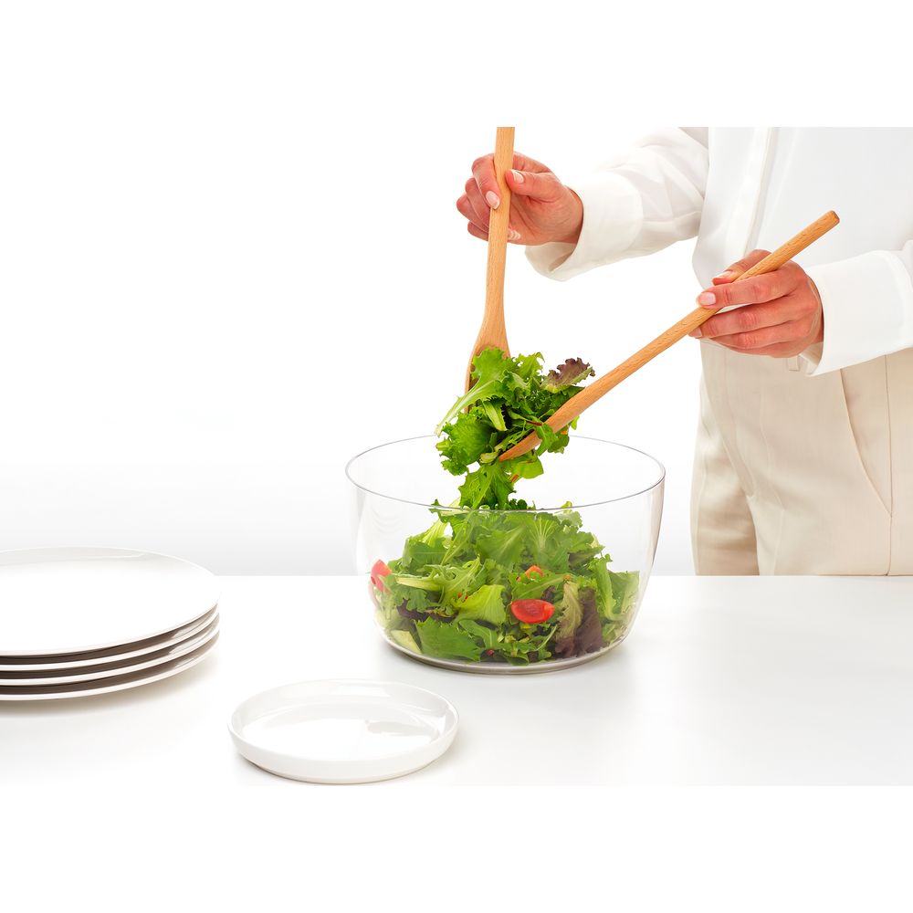 Mani che mescolano insalata verde con pomodori in una ciotola trasparente con cucchiai di legno. Sfondo bianco.