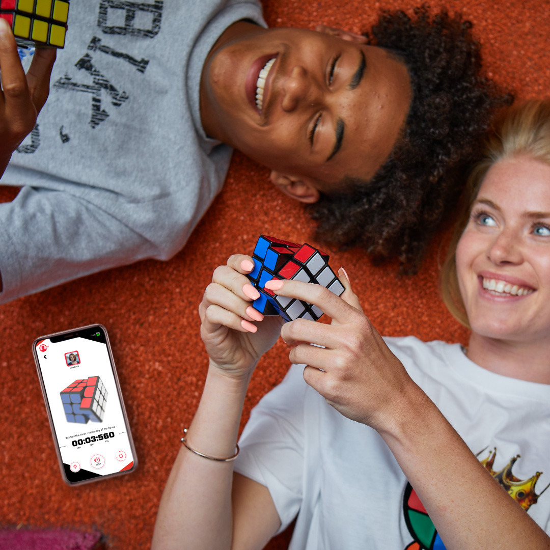 Due persone sorridenti giacciono su un tappeto arancione, giocando con i cubi di Rubik. Un telefono mostra un timer.