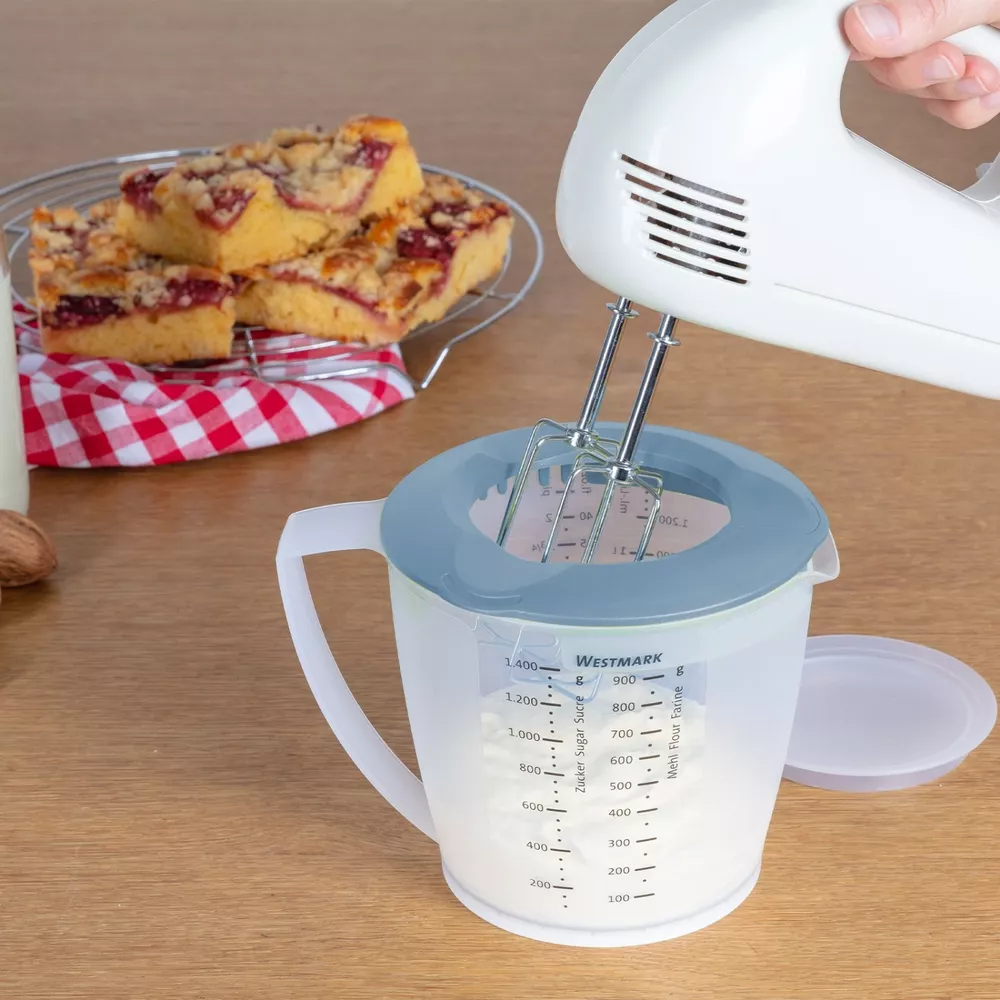 Ein Handmixer wird benutzt, um Mehl in einem Messbecher zu mischen. Kuchen liegt im Hintergrund.