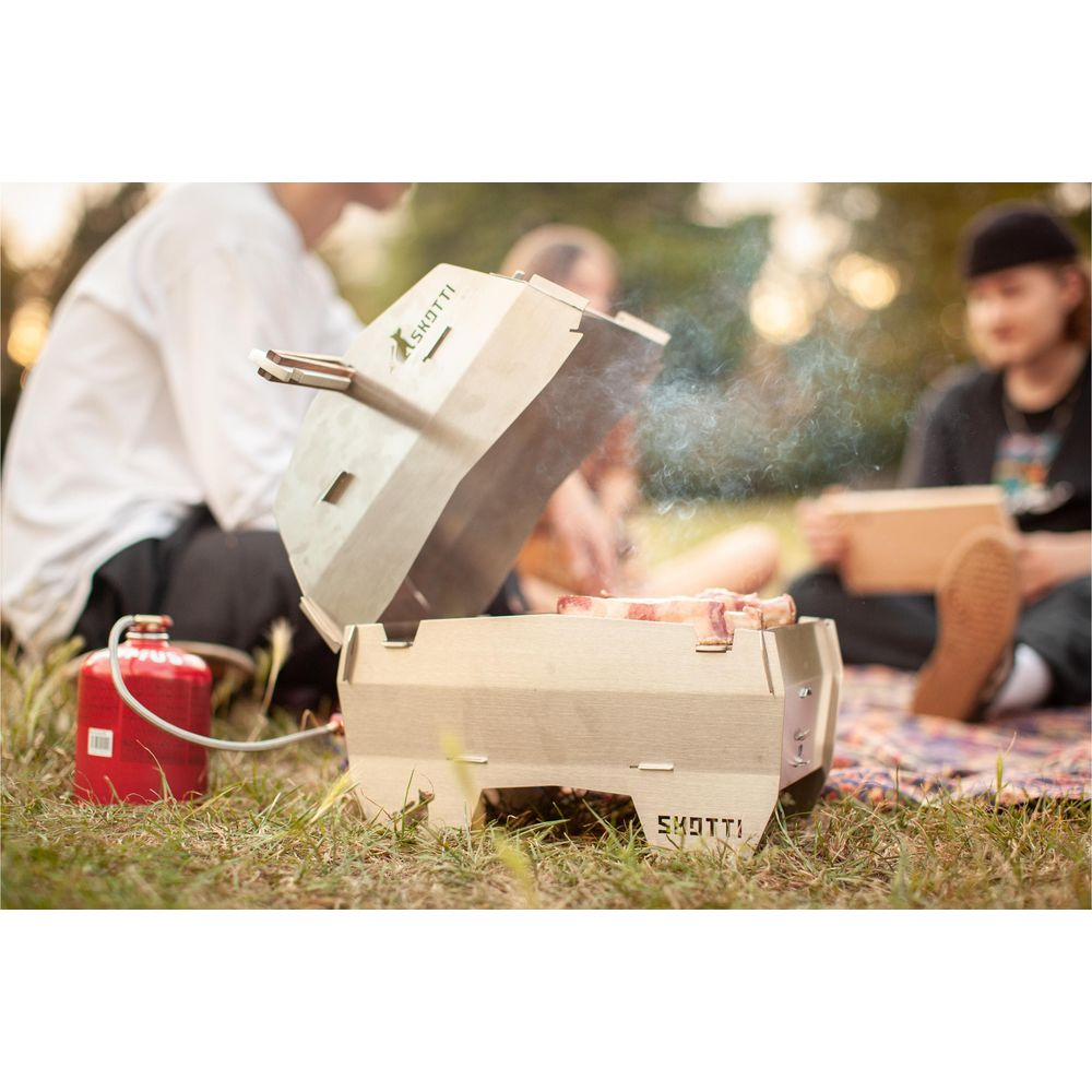 Una griglia portatile con carne che fuma all'aperto, persone sullo sfondo.