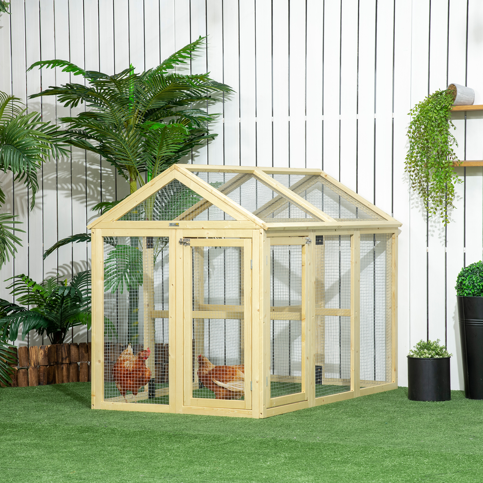 Un gallinero de madera con malla de alambre sobre césped artificial, con plantas y una pared blanca detrás.