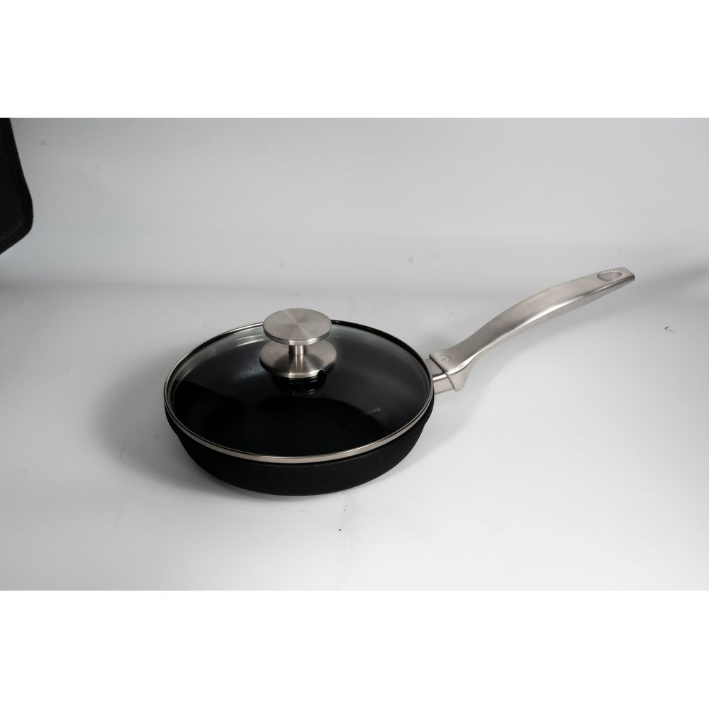 Une poêle noire avec couvercle en verre et manche argenté repose sur une surface blanche.