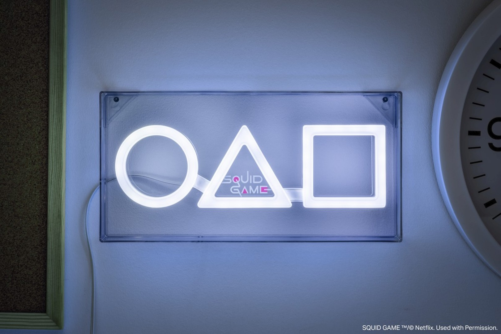Świecące logo "Squid Game" z kształtami koła, trójkąta i kwadratu na ścianie.