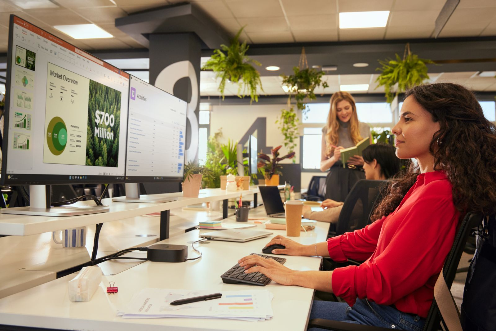Vrouw in rood shirt typt aan bureau met dubbele monitor. Anderen werken op achtergrond.