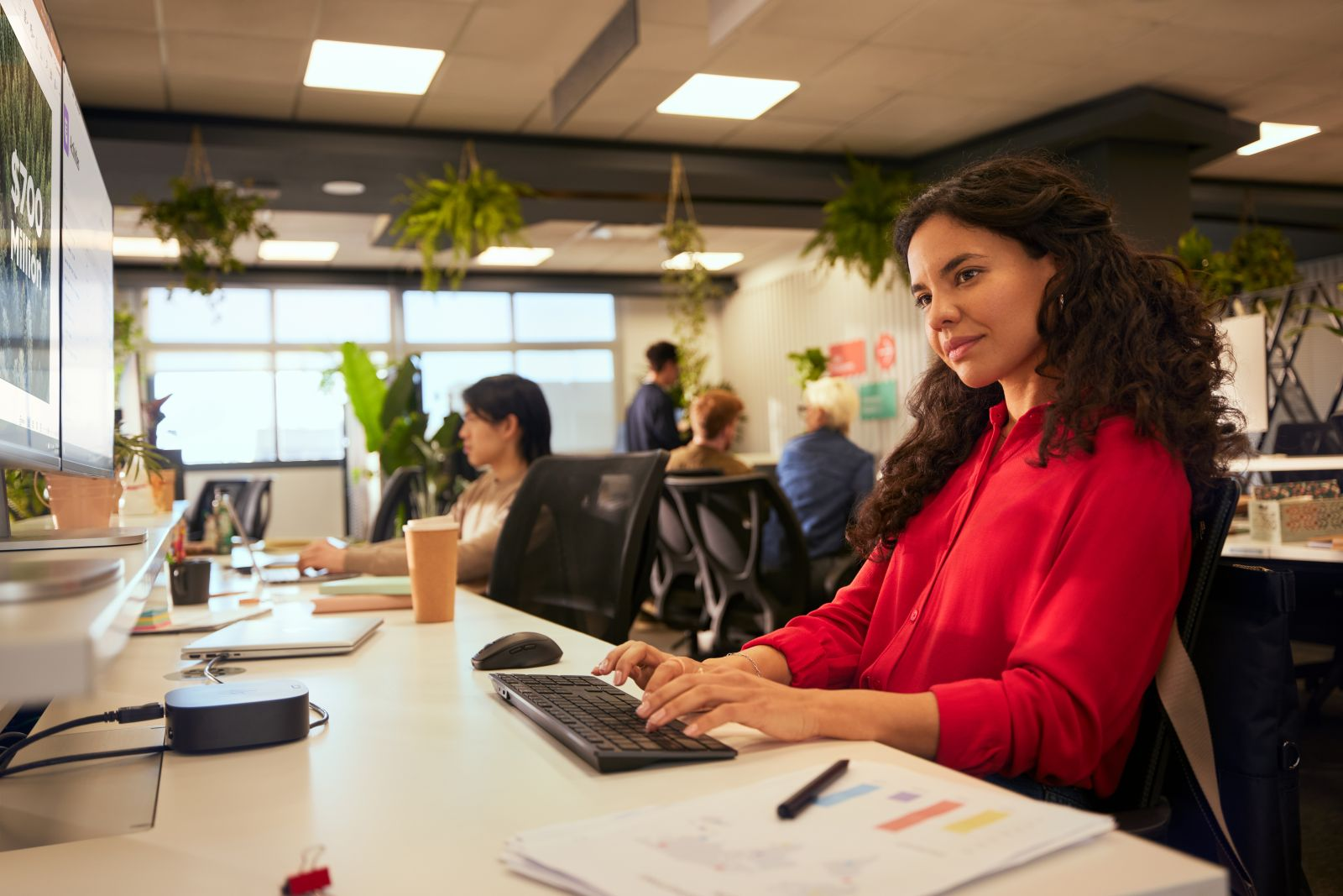Vrouw in rood shirt typt op toetsenbord in lichte kantoor met planten en collega's.