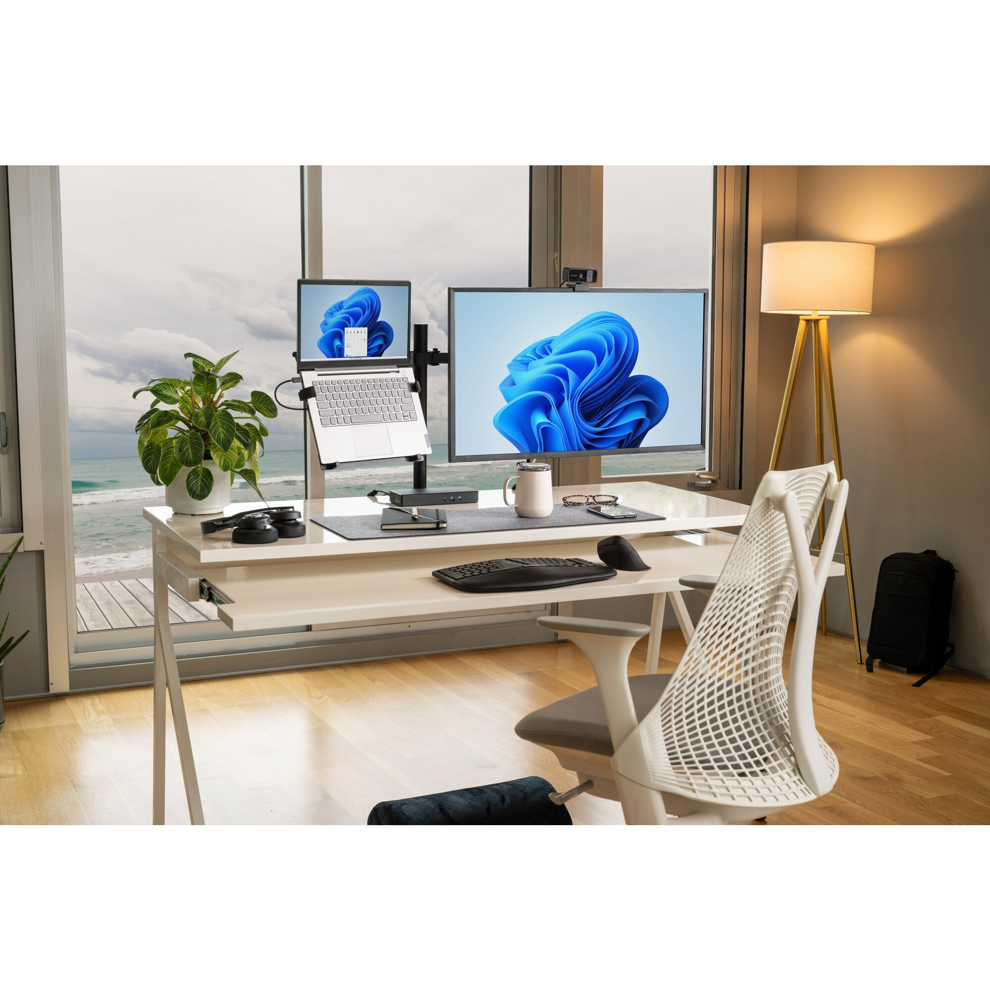 A desk with dual monitors and a laptop in front of a window overlooking the sea.