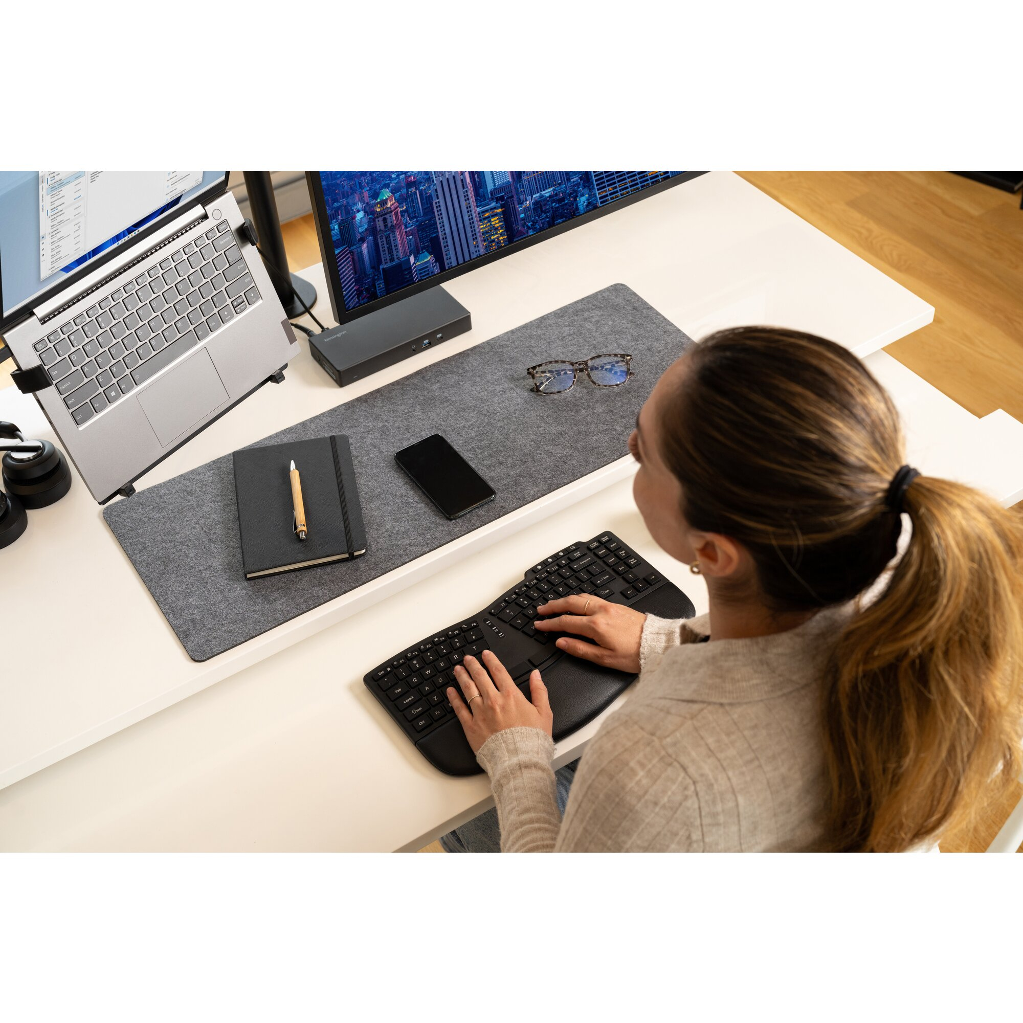 Mujer tecleando en teclado, portátil, monitor y libreta en escritorio.