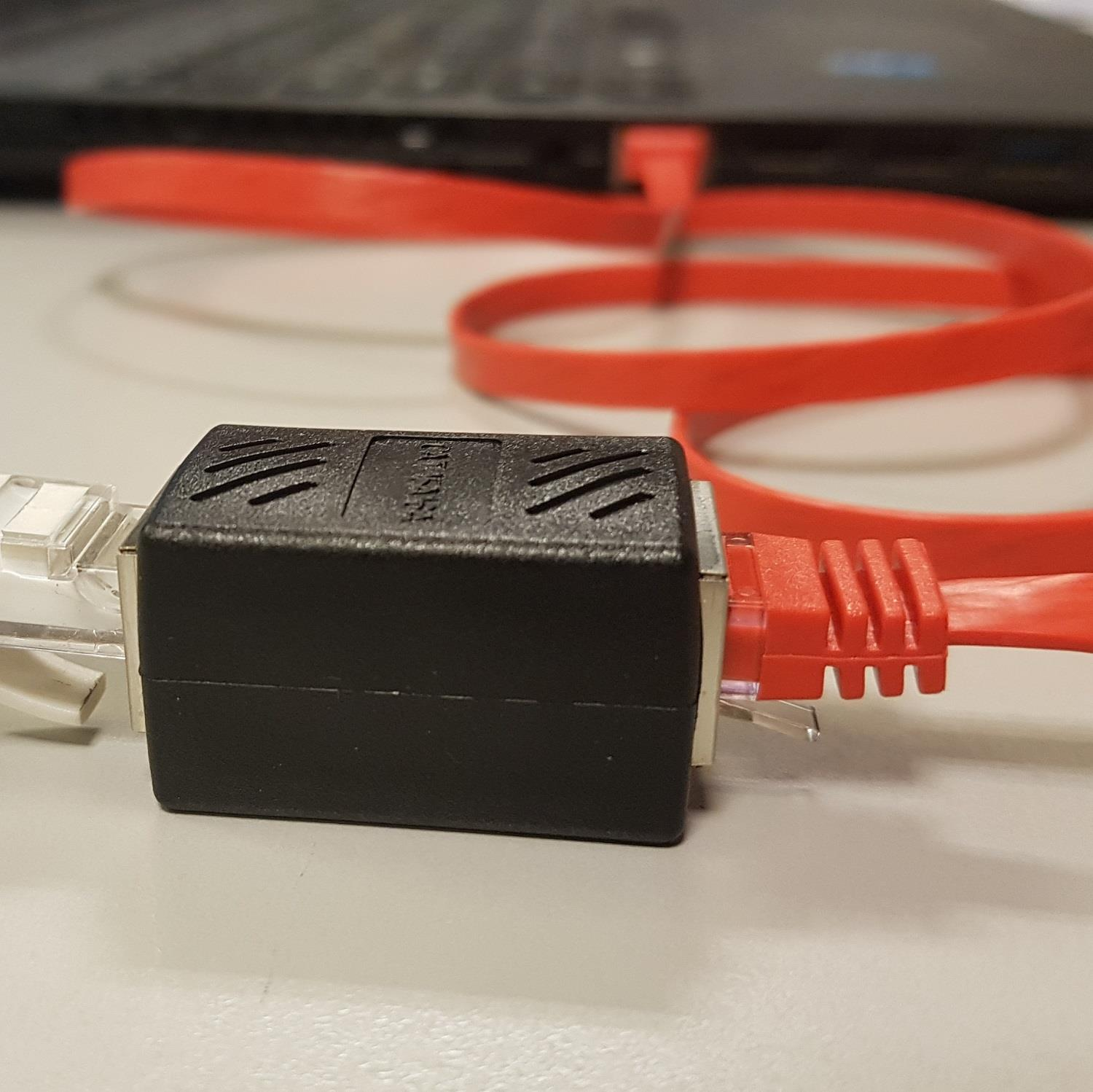 Siyah bir adaptör iki kırmızı ağ kablosunu bağlıyor. Arkada bir dizüstü bilgisayar.