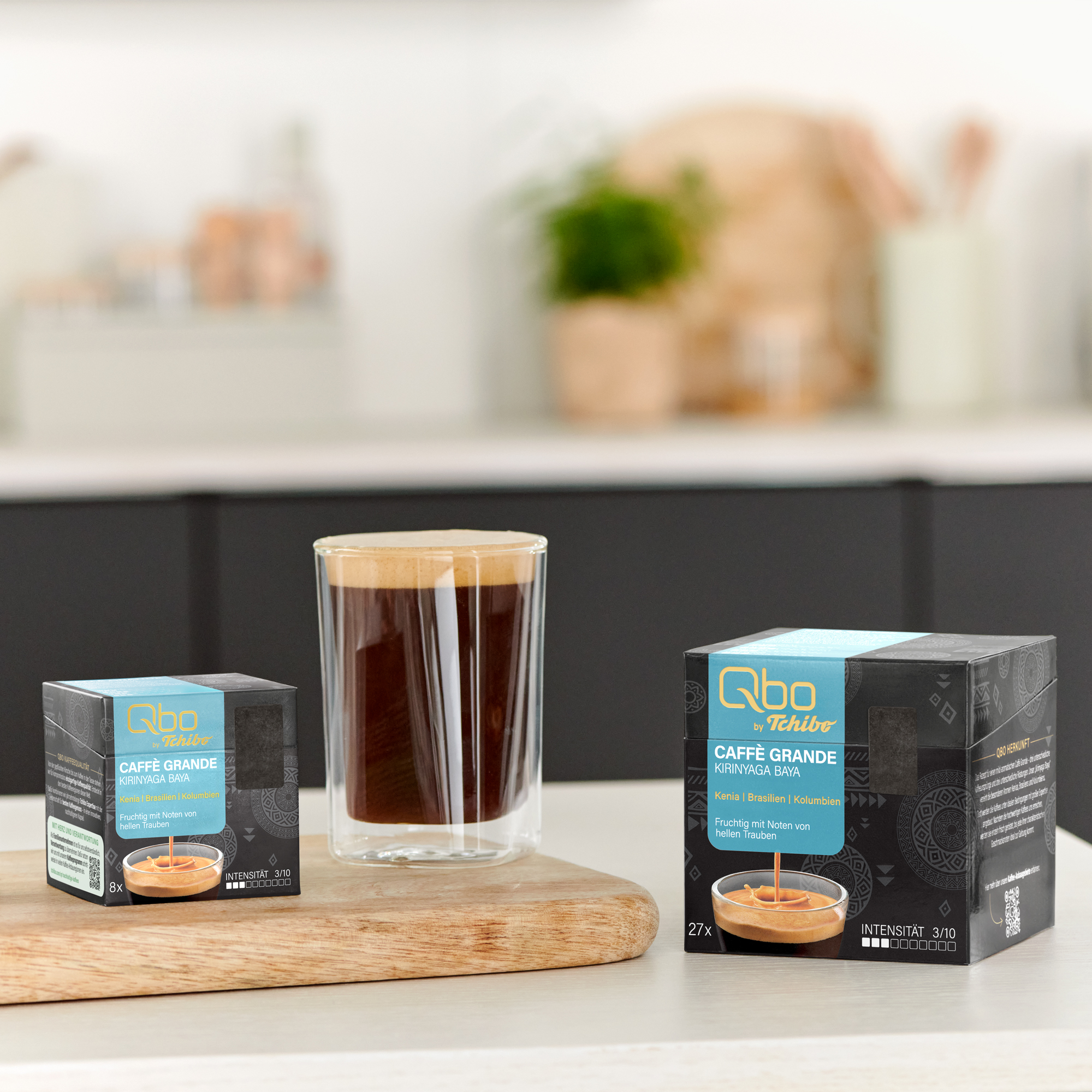 Zwei Packungen Tchibo Qbo Kaffee und ein Glas Kaffee mit Schaum auf einem Holzteller.