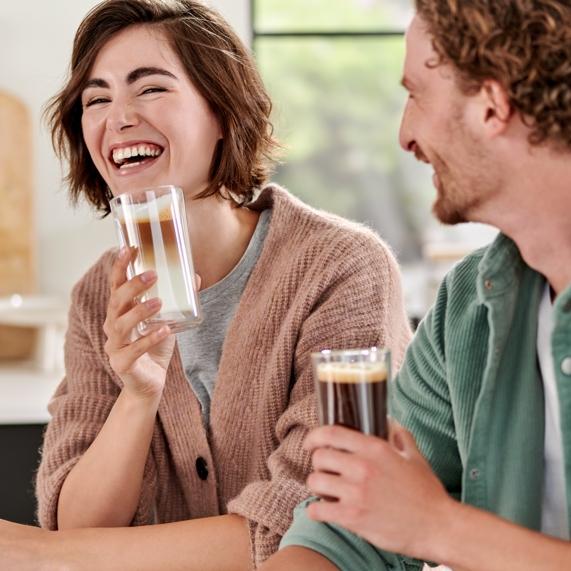 Ein Mann und eine Frau lachen und halten Getränke in einem hellen Raum.
