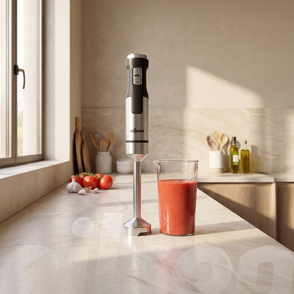 Una batidora de inmersión plateada junto a un vaso de batido rojo sobre una encimera de mármol.