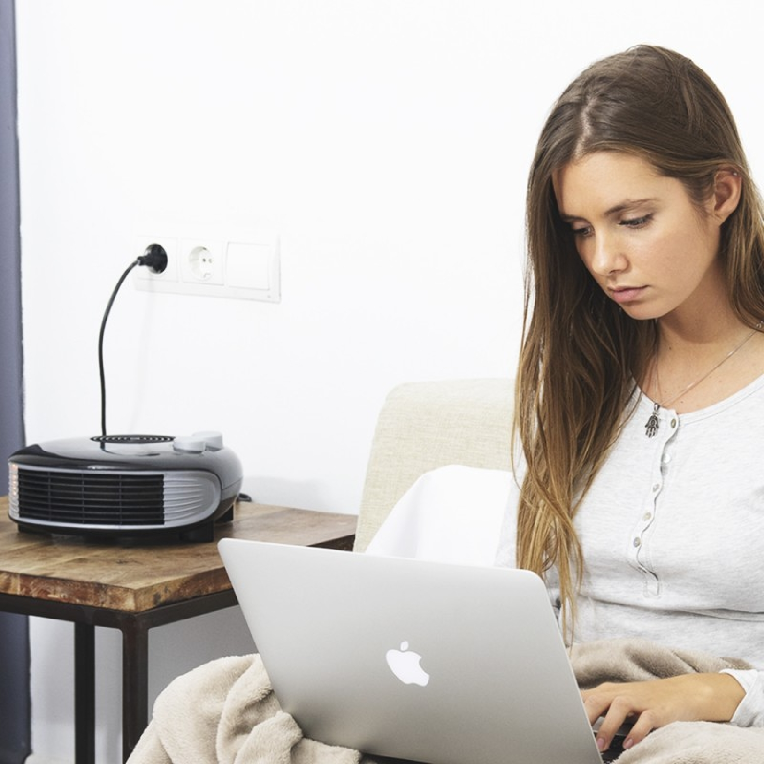 Een vrouw zit op een bank en gebruikt een laptop. Een kachel staat ernaast op een tafel.