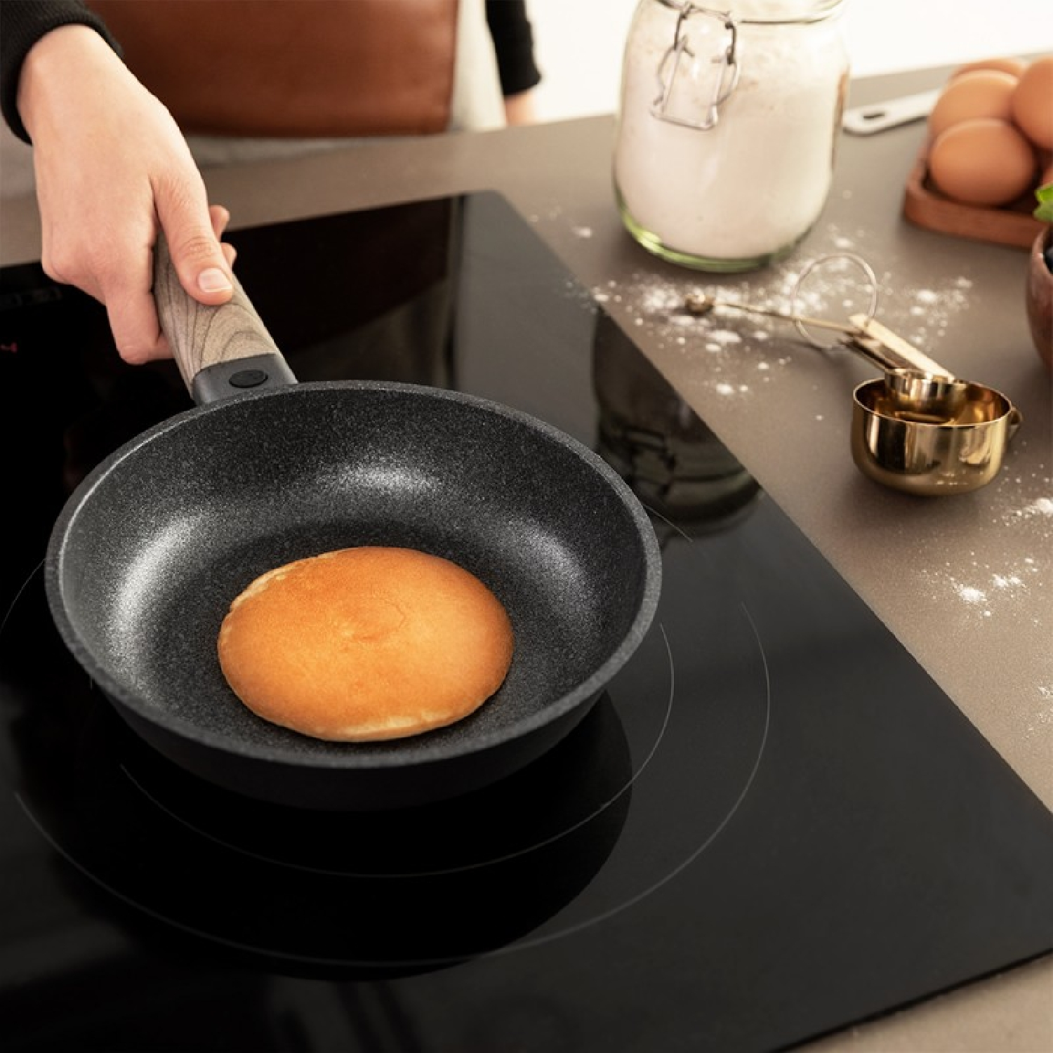 Una mano tiene una padella con un pancake dorato, su un piano cottura a induzione nero.